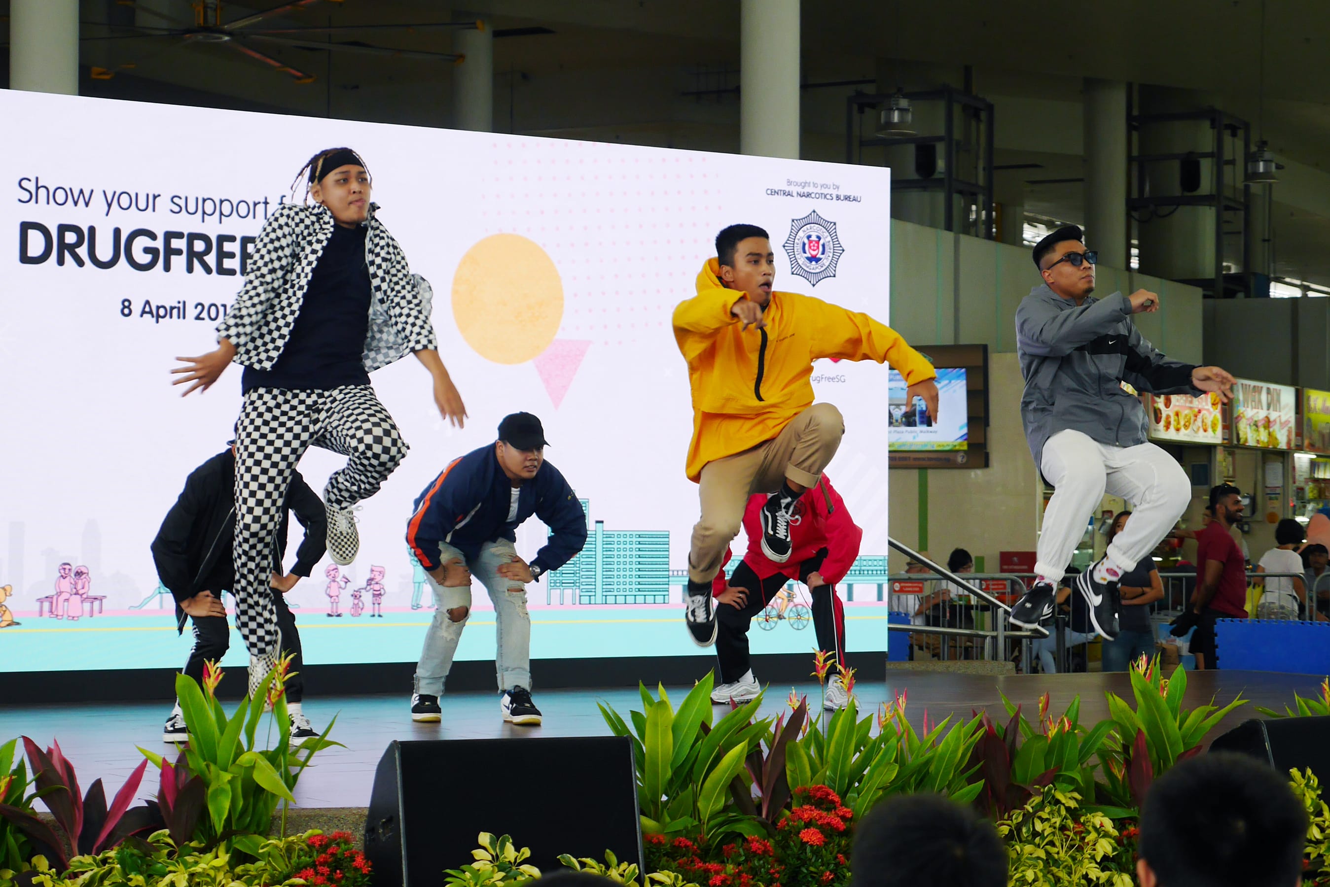Six dancers on stage jumping, with a "DrugFree" backdrop and potted plants in the foreground.