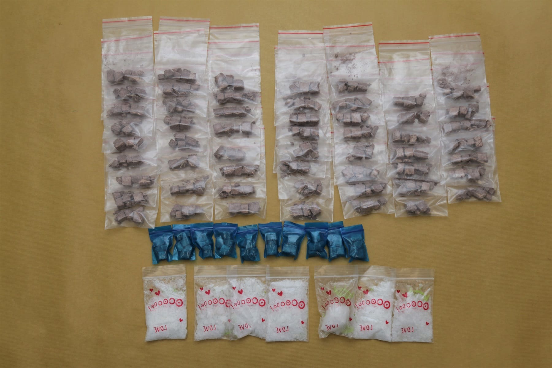 Rows of plastic bags containing chunks, blue pills, and white powder on a neutral surface.
