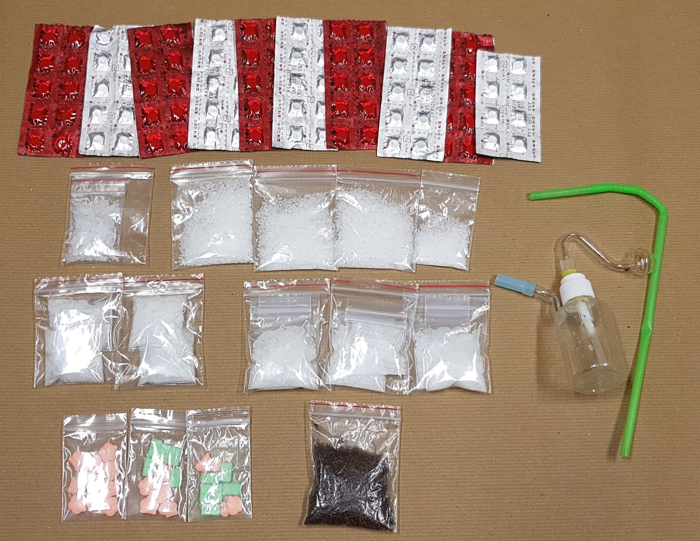 Blister packs, bags of crystals, bear pills, dark flakes, and a glass pipe are arranged on a brown surface.