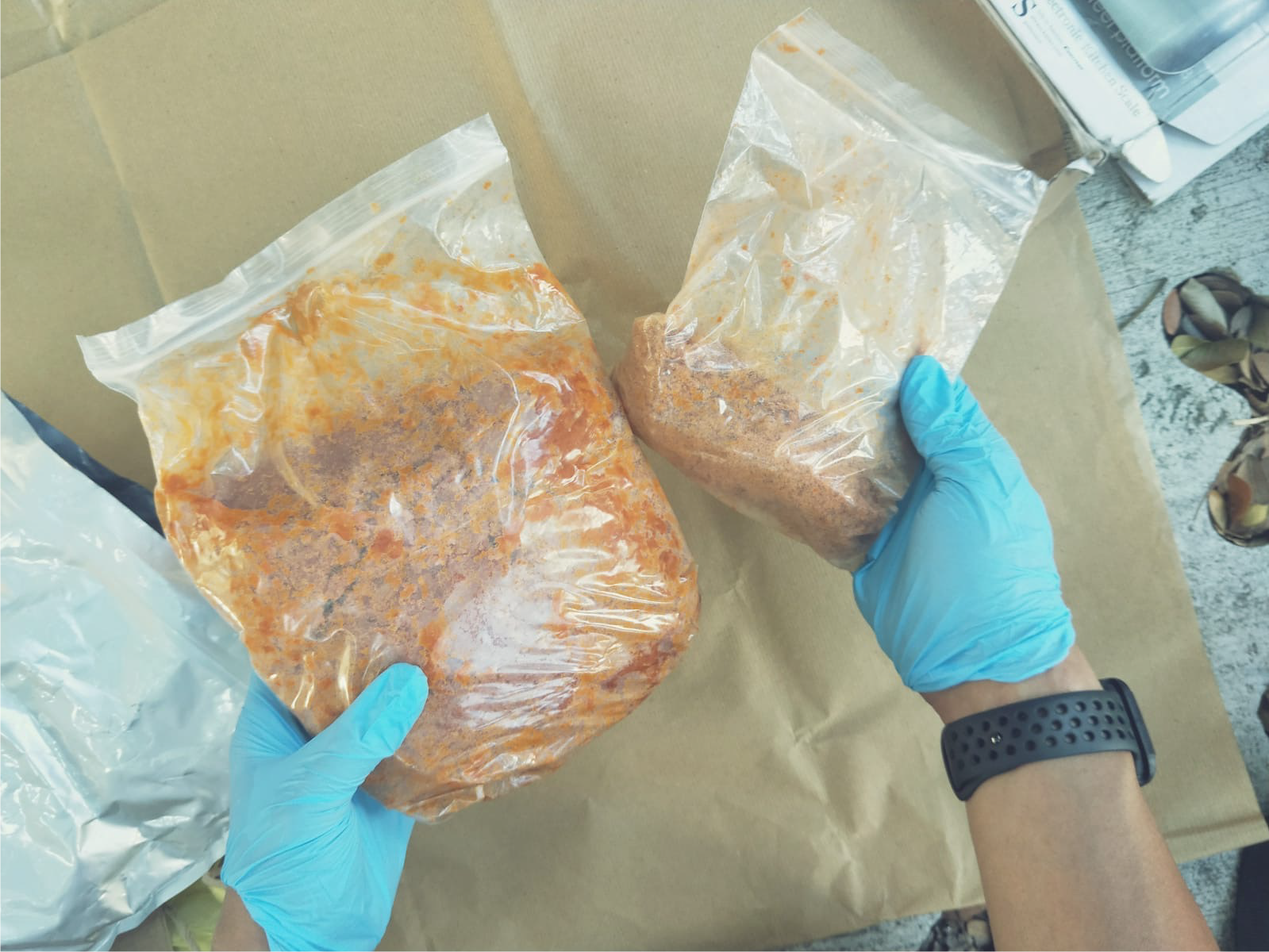 Two gloved hands hold plastic bags with orange/brown food. Kraft paper background.