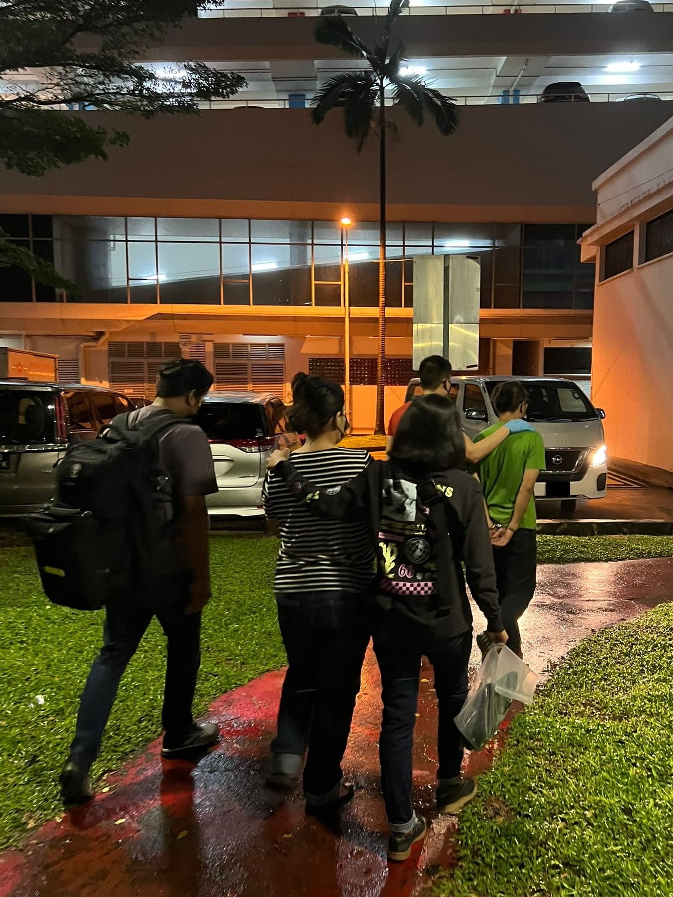 Group walking on wet pavement toward building; one person appears handcuffed, escorted by another.