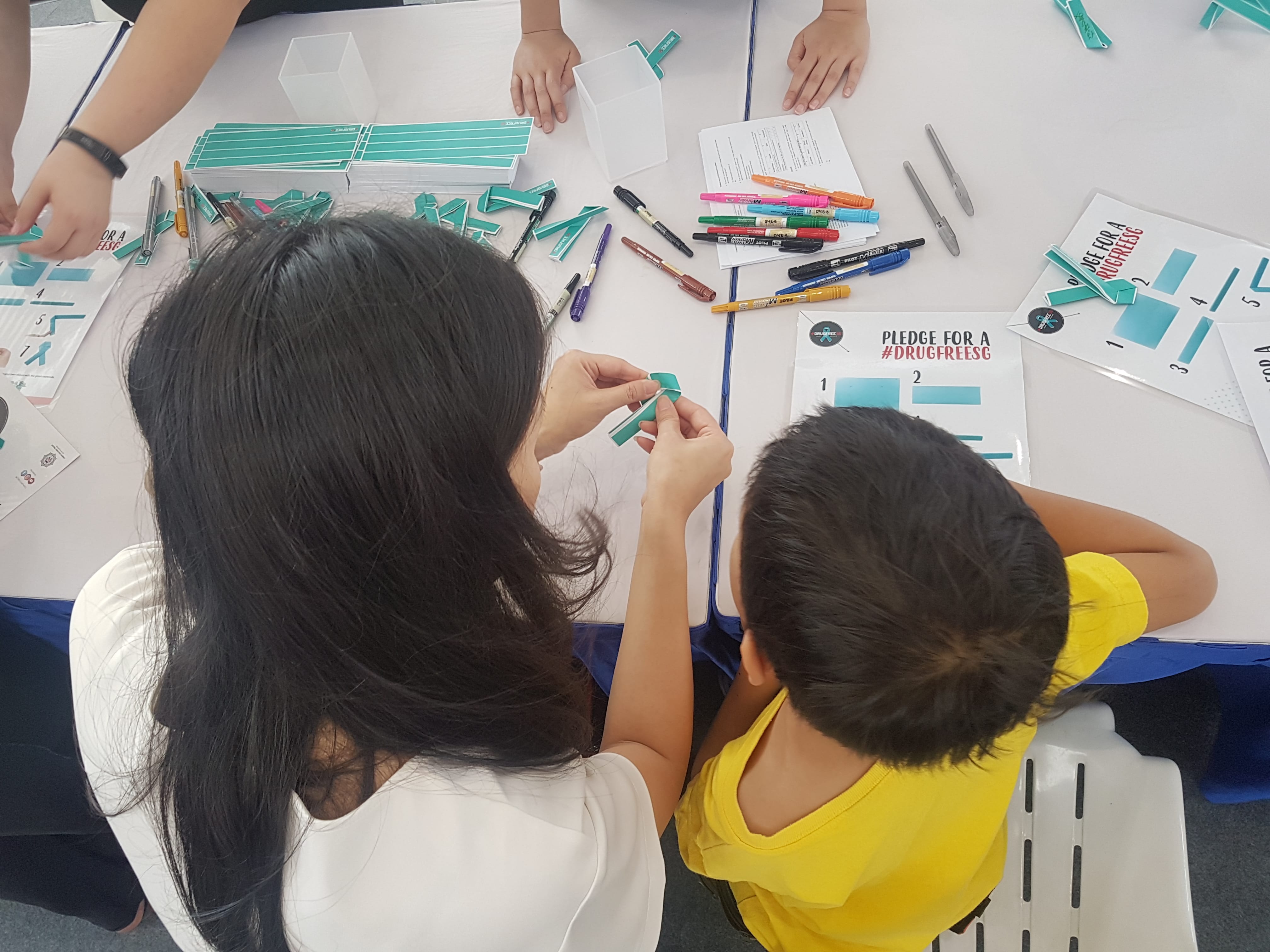 Overhead view: People assemble #DrugFreeSG pledge ribbons at a white table covered in pens and instruction sheets.