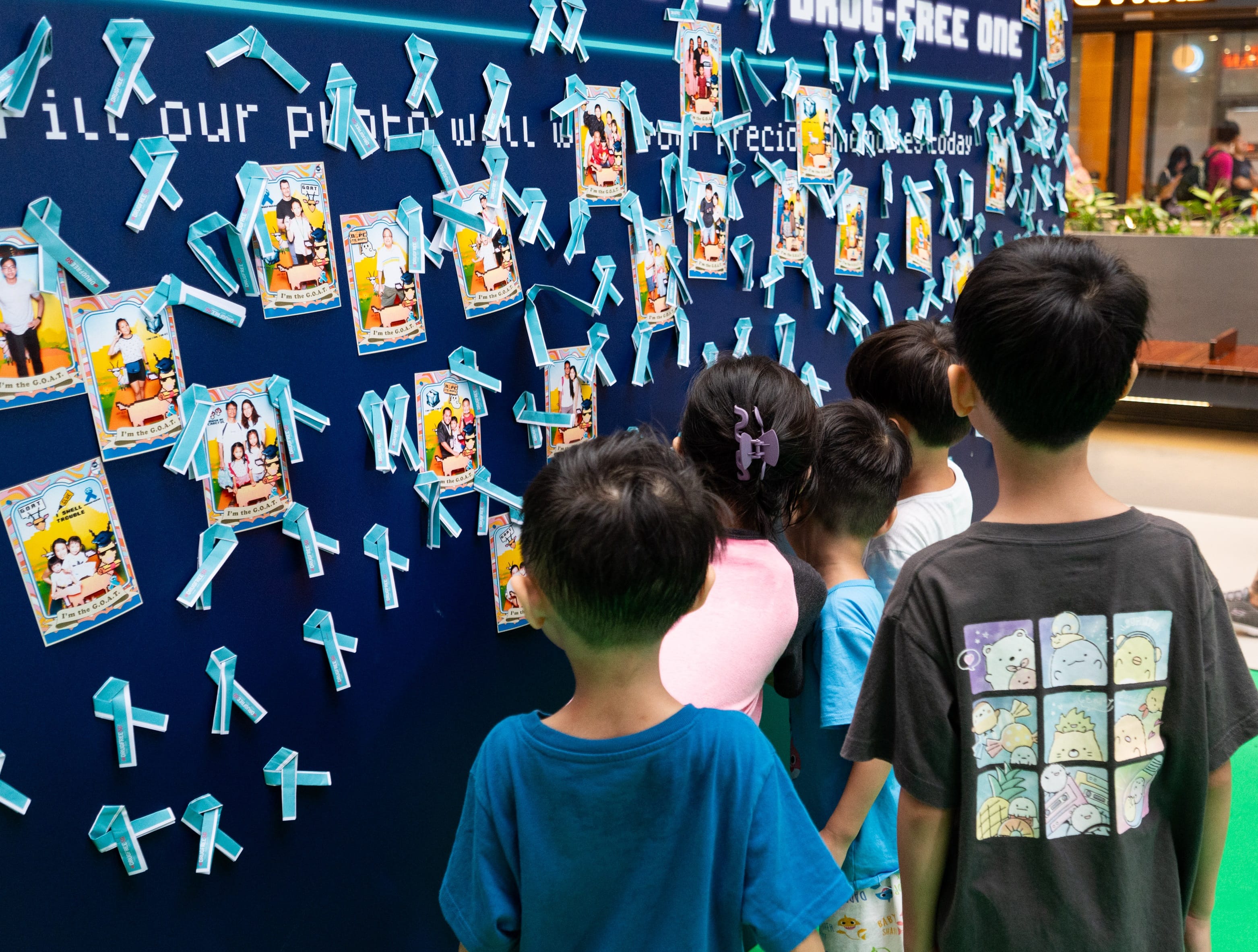 Children face a blue wall with family photos & blue ribbons promoting "Drug-Free." Boy wears "Sumikko Gurashi" character tee.