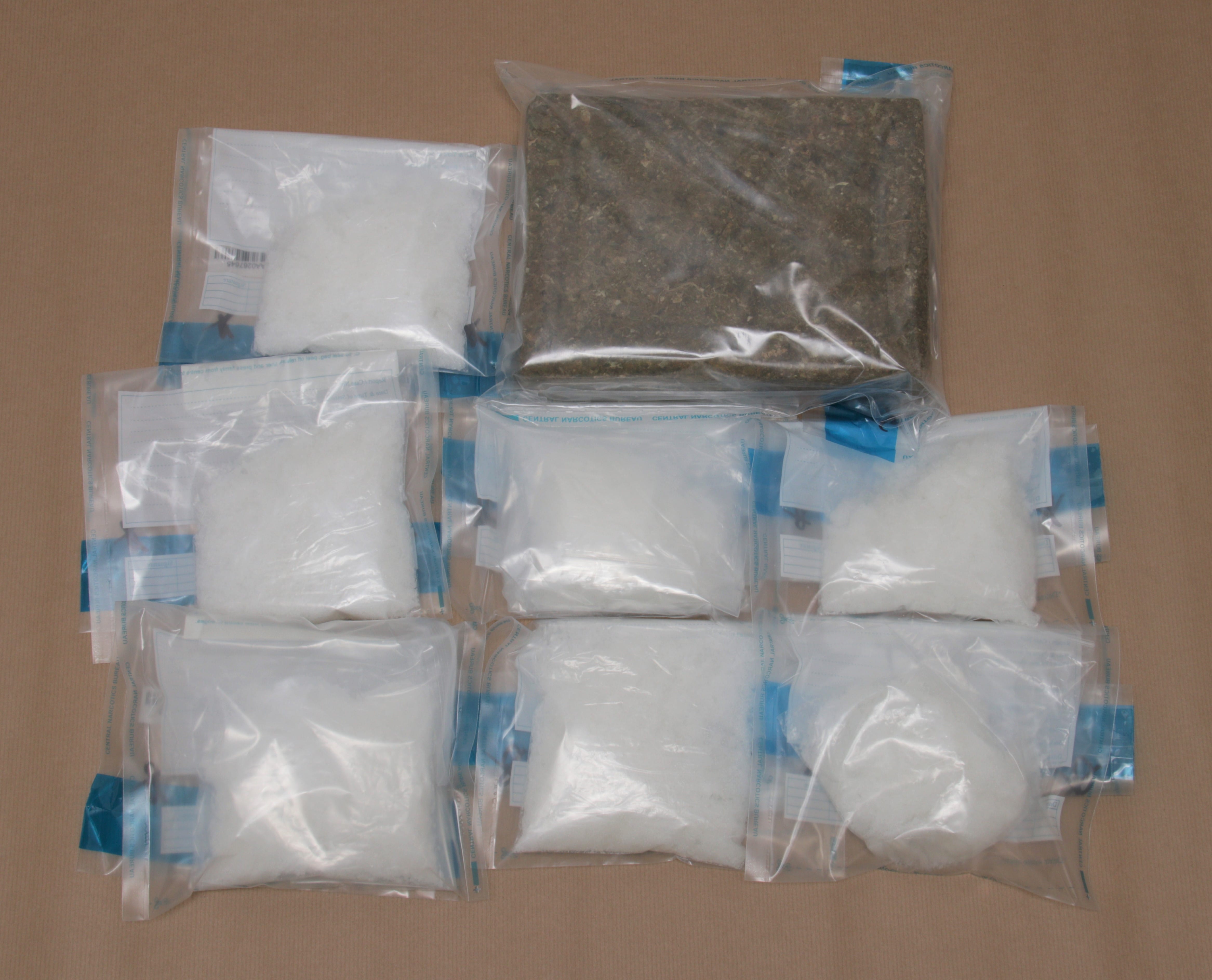 Eight clear bags on a brown surface; seven filled with white powder, one with a green leafy substance.