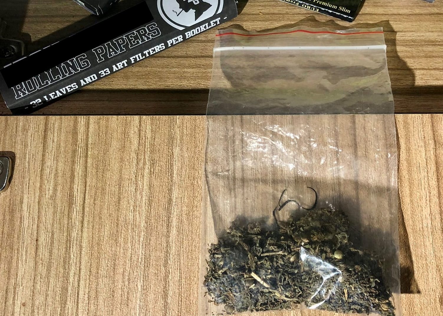 Rolling Papers pack beside a plastic bag with dried leaves on a wooden surface.