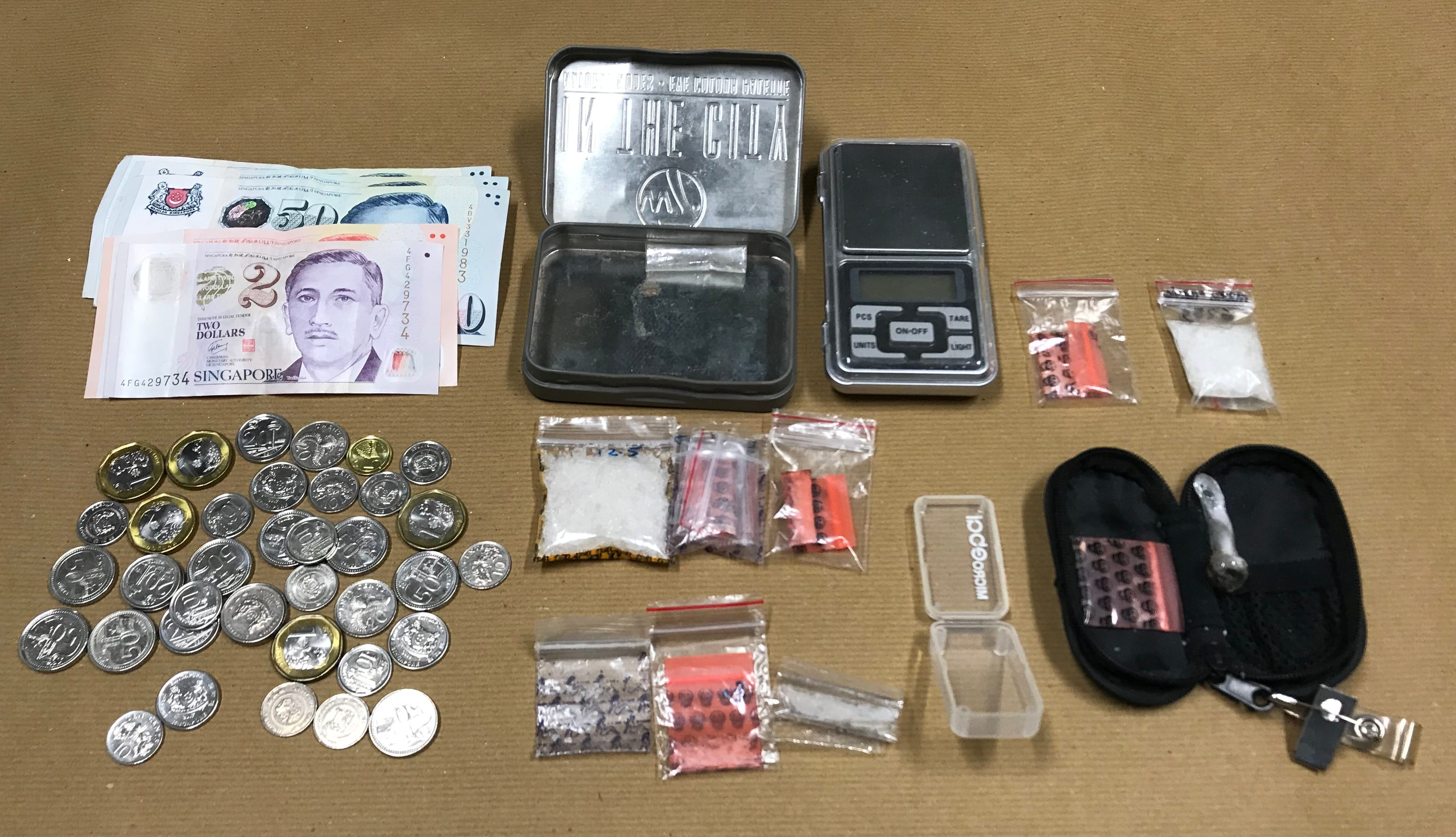 Singapore money, coins, digital scale, small baggies with pills and powder, a pipe, and a metal container.