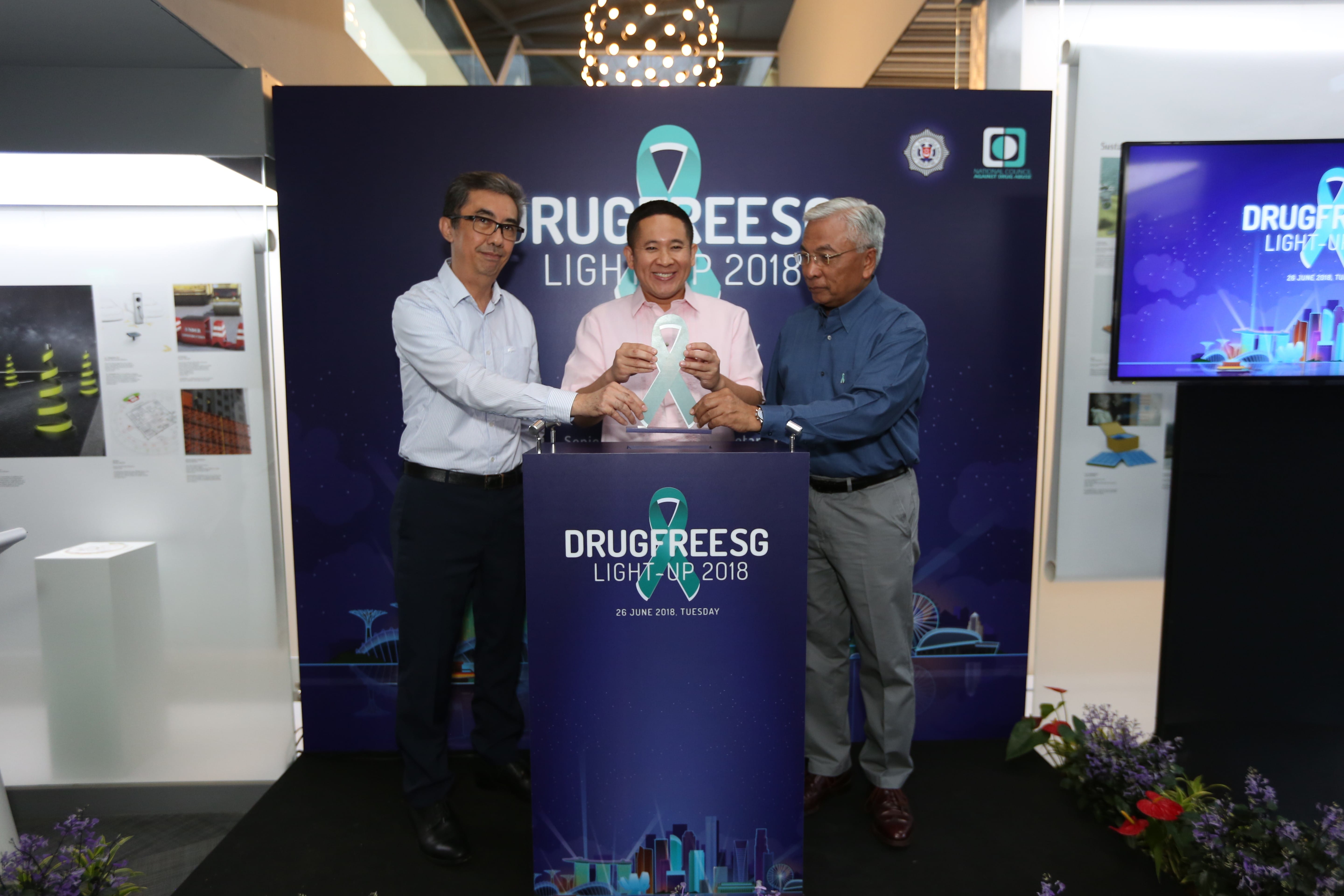Three men holding a teal ribbon shape at a DrugFreeSG event stand with a backdrop.
