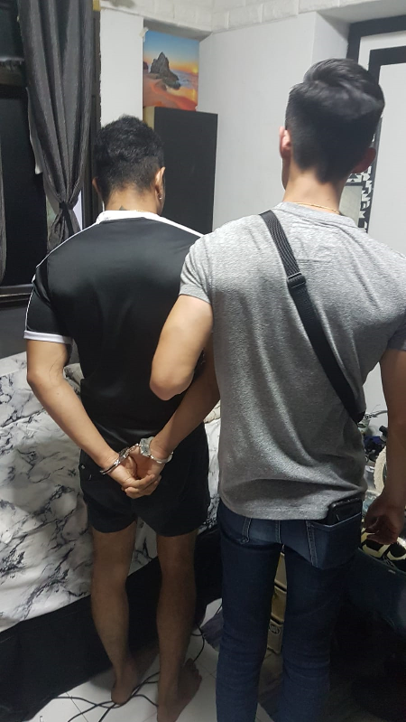 Two people standing; one handcuffed with hands behind back, other with shoulder bag.