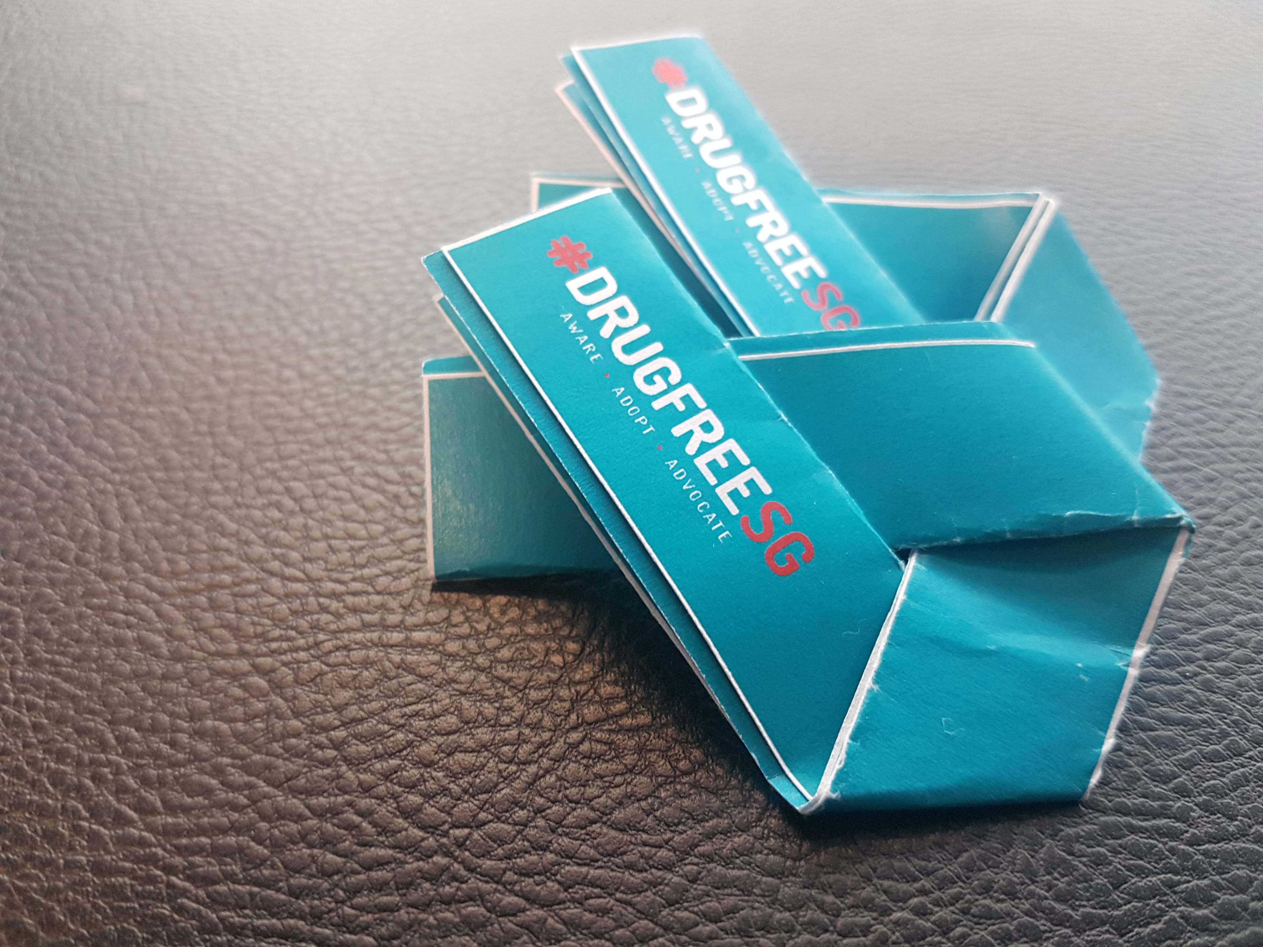 Teal "DrugFreeSG" awareness ribbons are stacked on a textured dark surface.