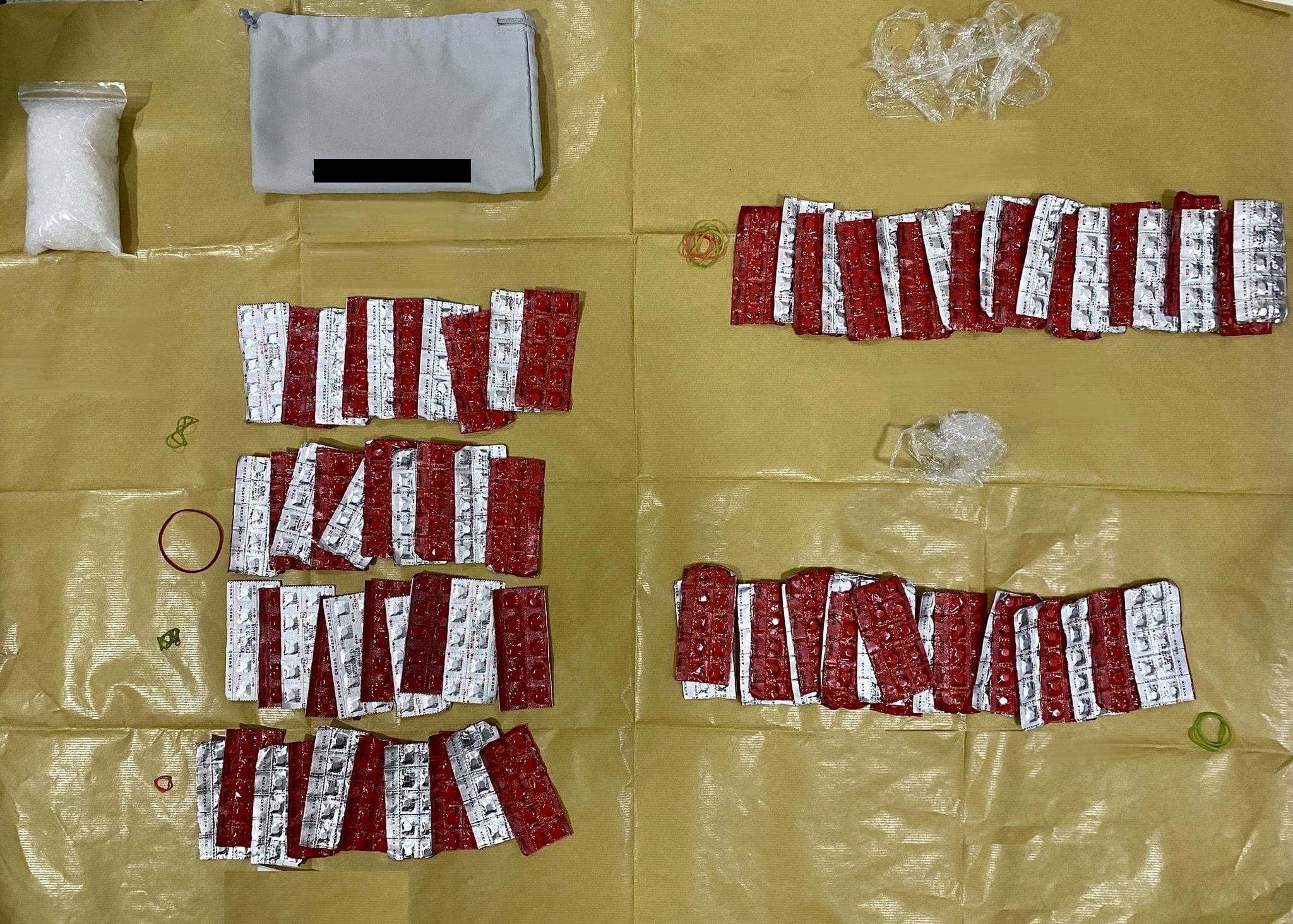 Red and silver pills in blister packs, clear plastic bag, gray pouch, rubber bands on tan surface.