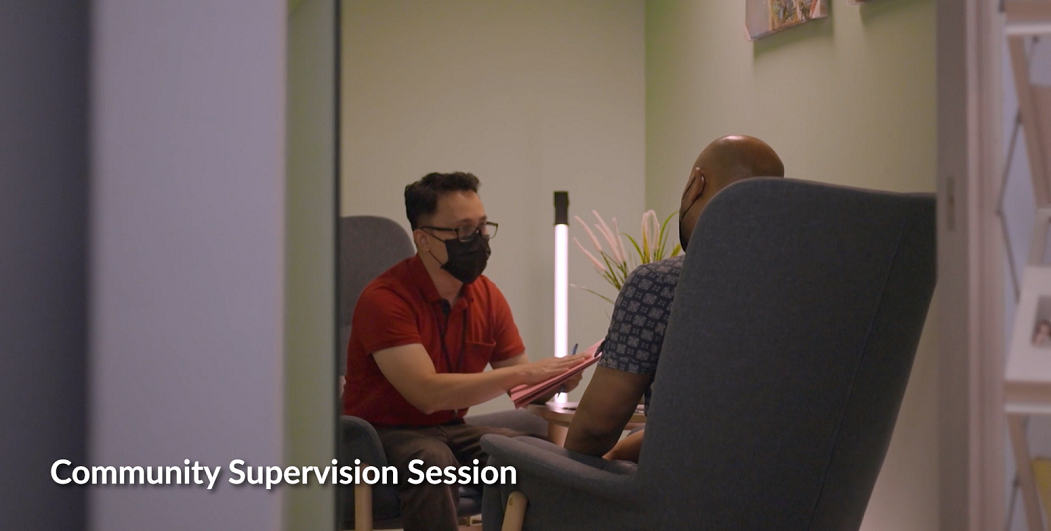 Two masked men seated in chairs; one man holding a clipboard, the other listening.