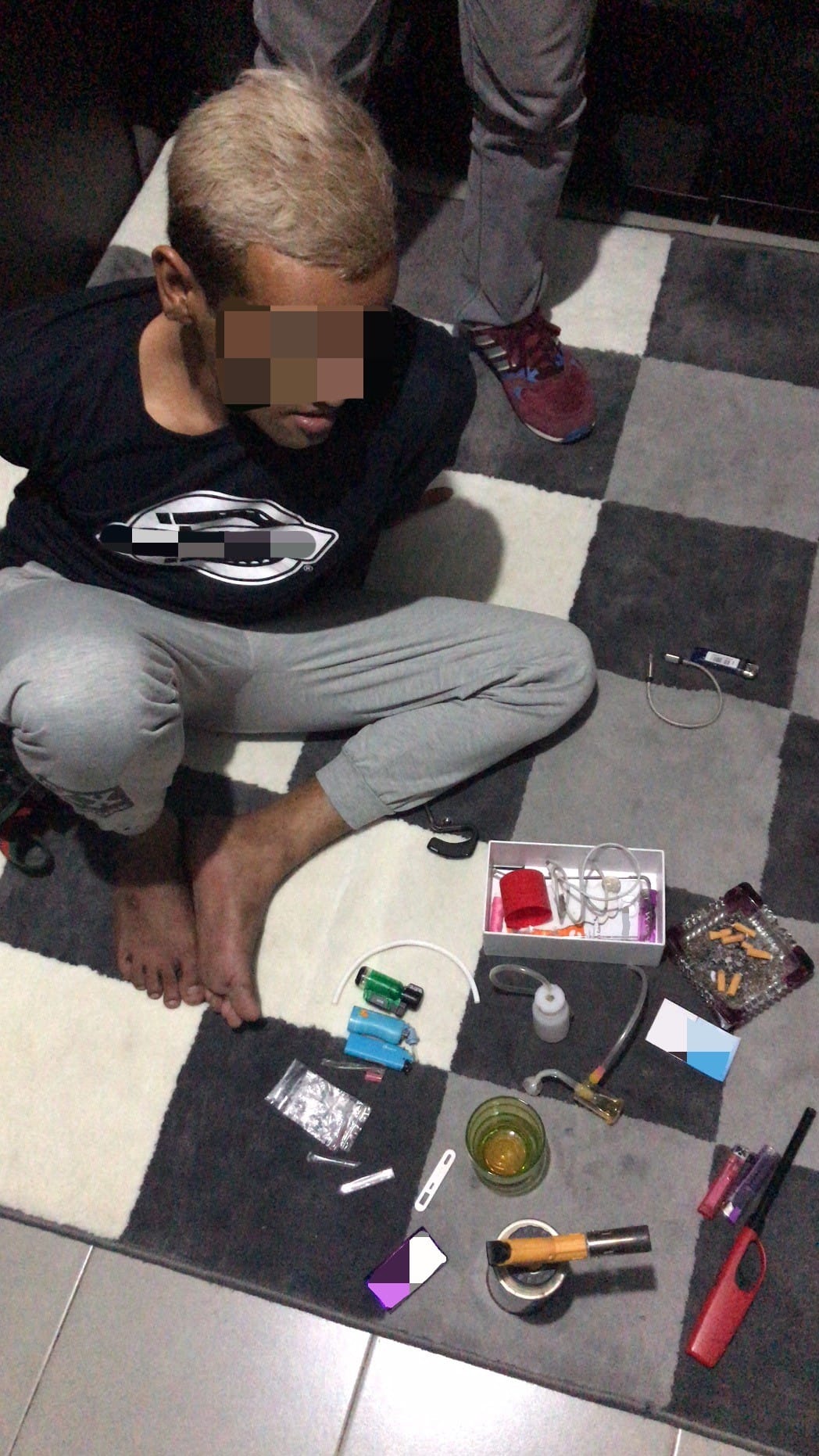 Person with blurred face squats on checkered carpet amongst drug paraphernalia. Another person stands behind.