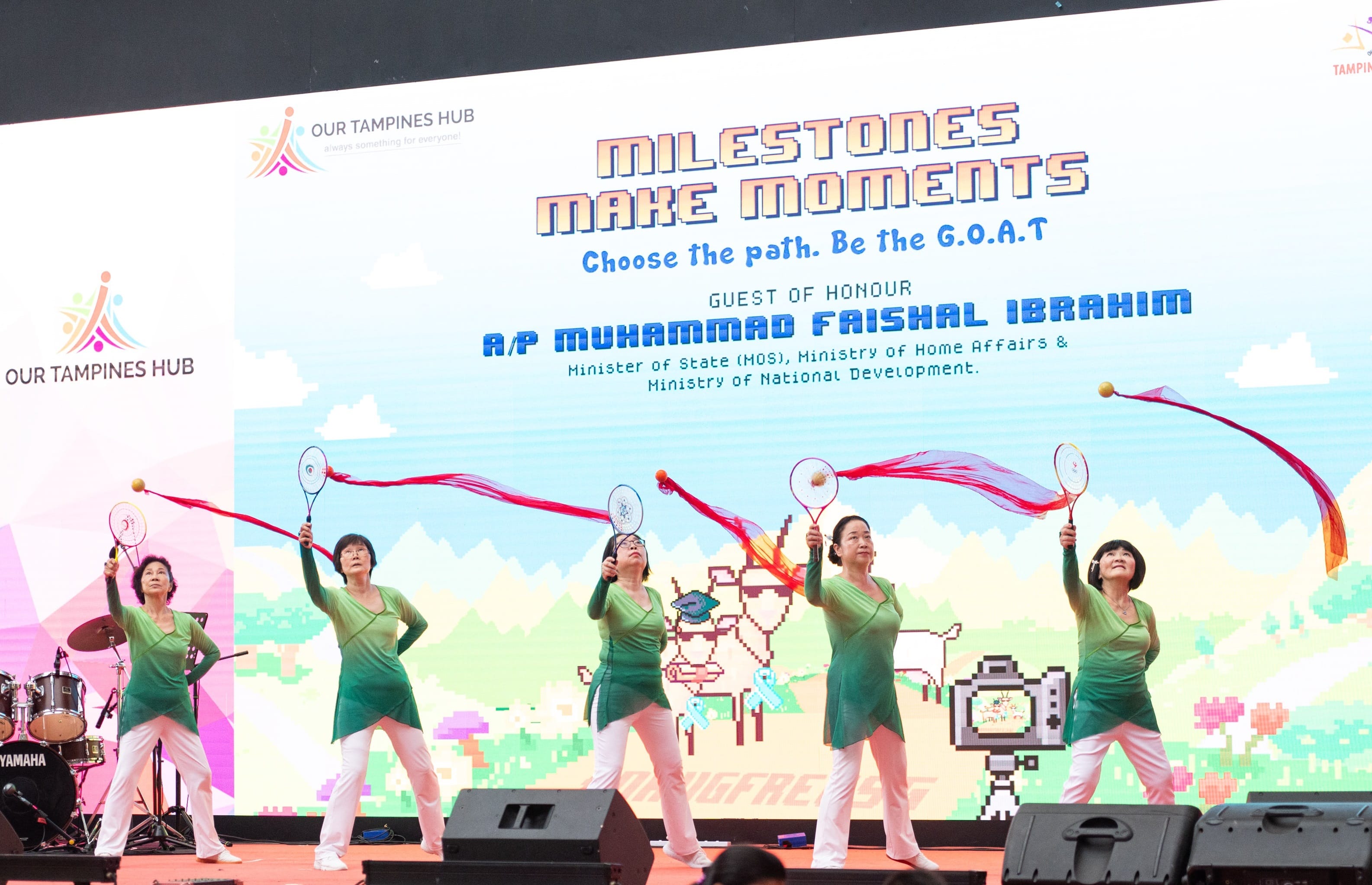 Five women in matching outfits perform with badminton rackets and red streamers on a stage.