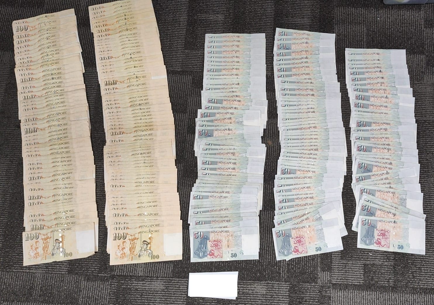 Stacks of Singapore $100 and $50 notes on a dark surface with a small white card.