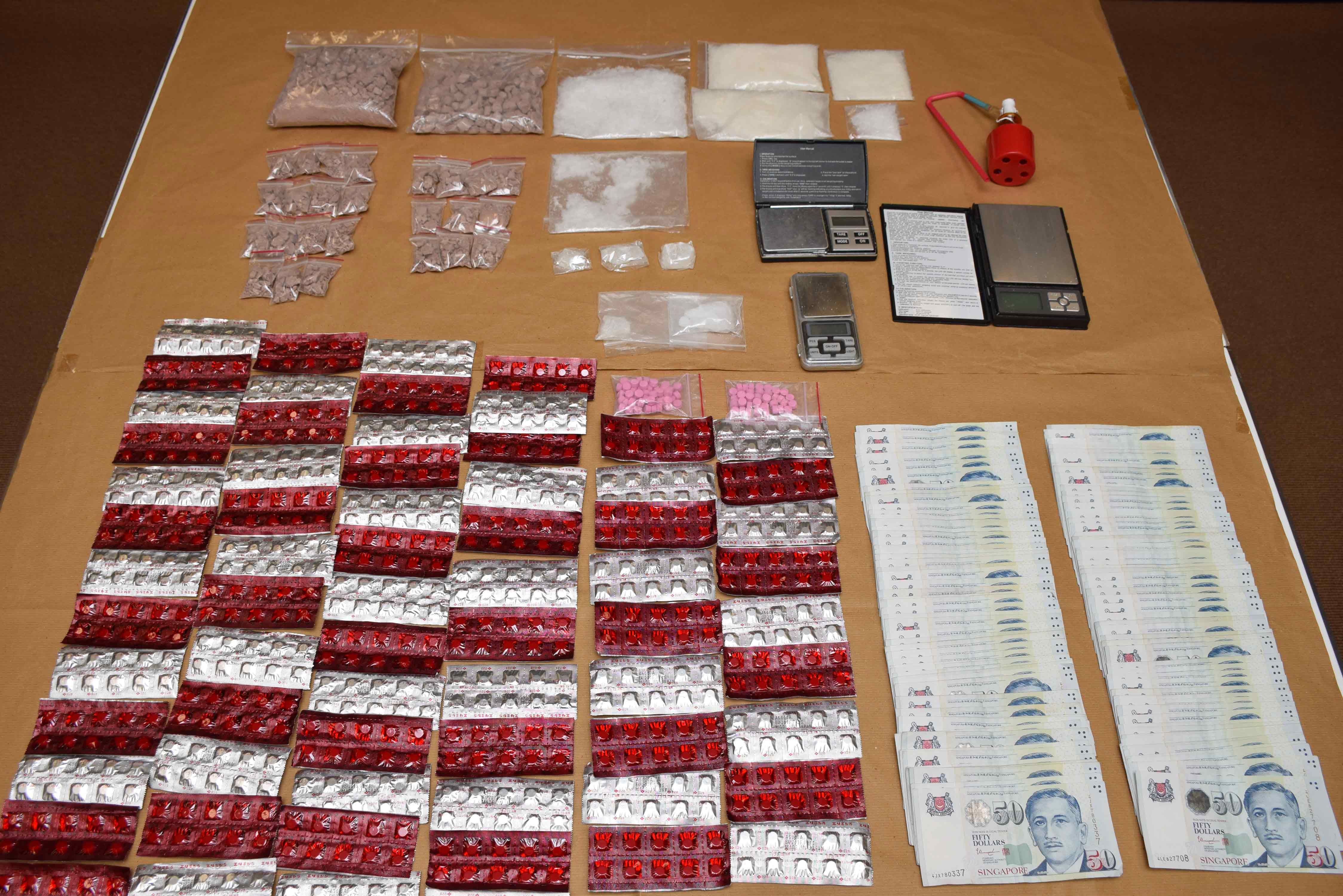 Drugs in bags and blister packs, scales, a red bottle, and stacks of Singapore fifty-dollar bills.