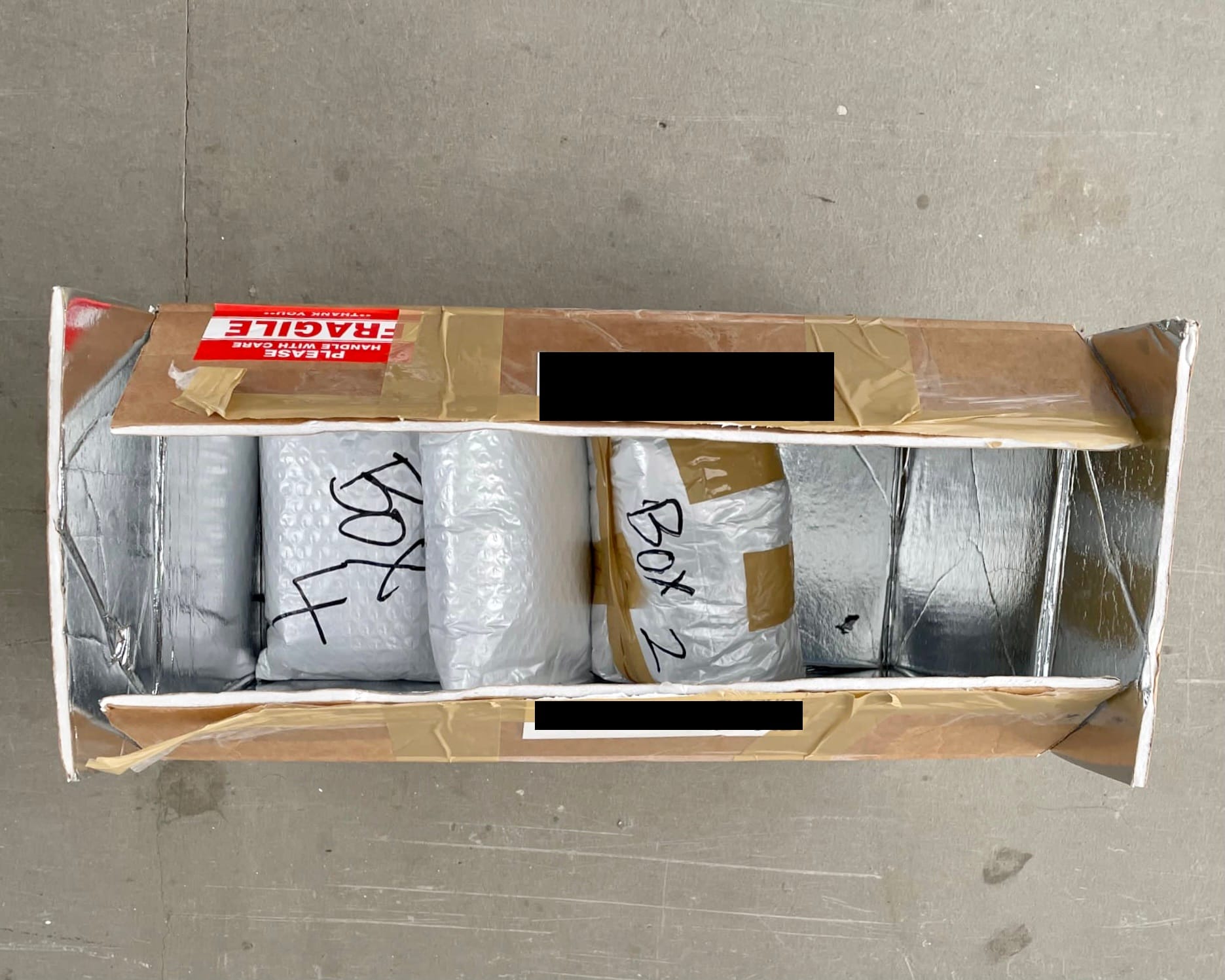 Open cardboard box with "Fragile" label, foil lining, and sealed white bags labeled "Box 1" and "Box 2".