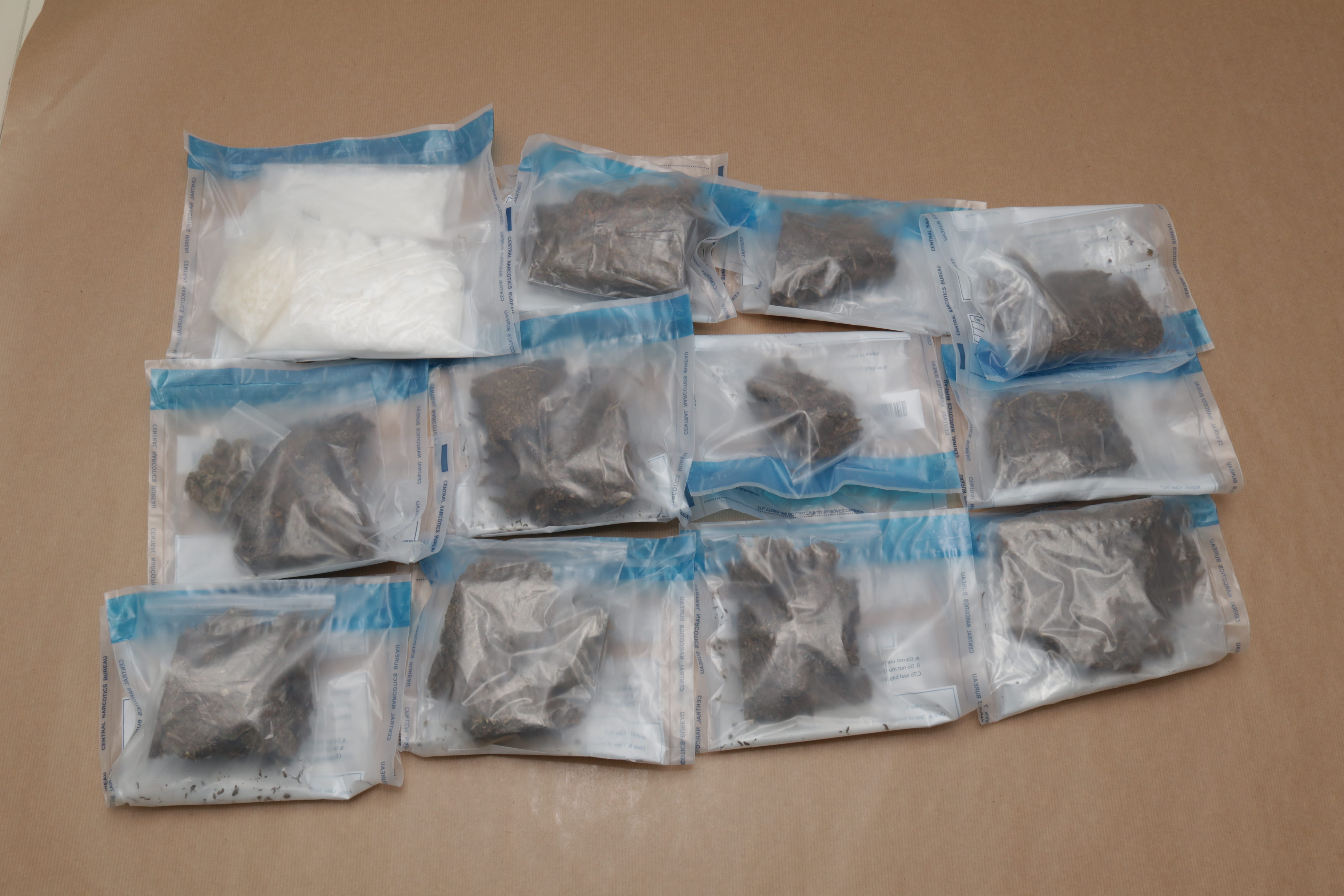 Eleven clear plastic bags, one with white powder, ten with brown leafy material, against a brown background.