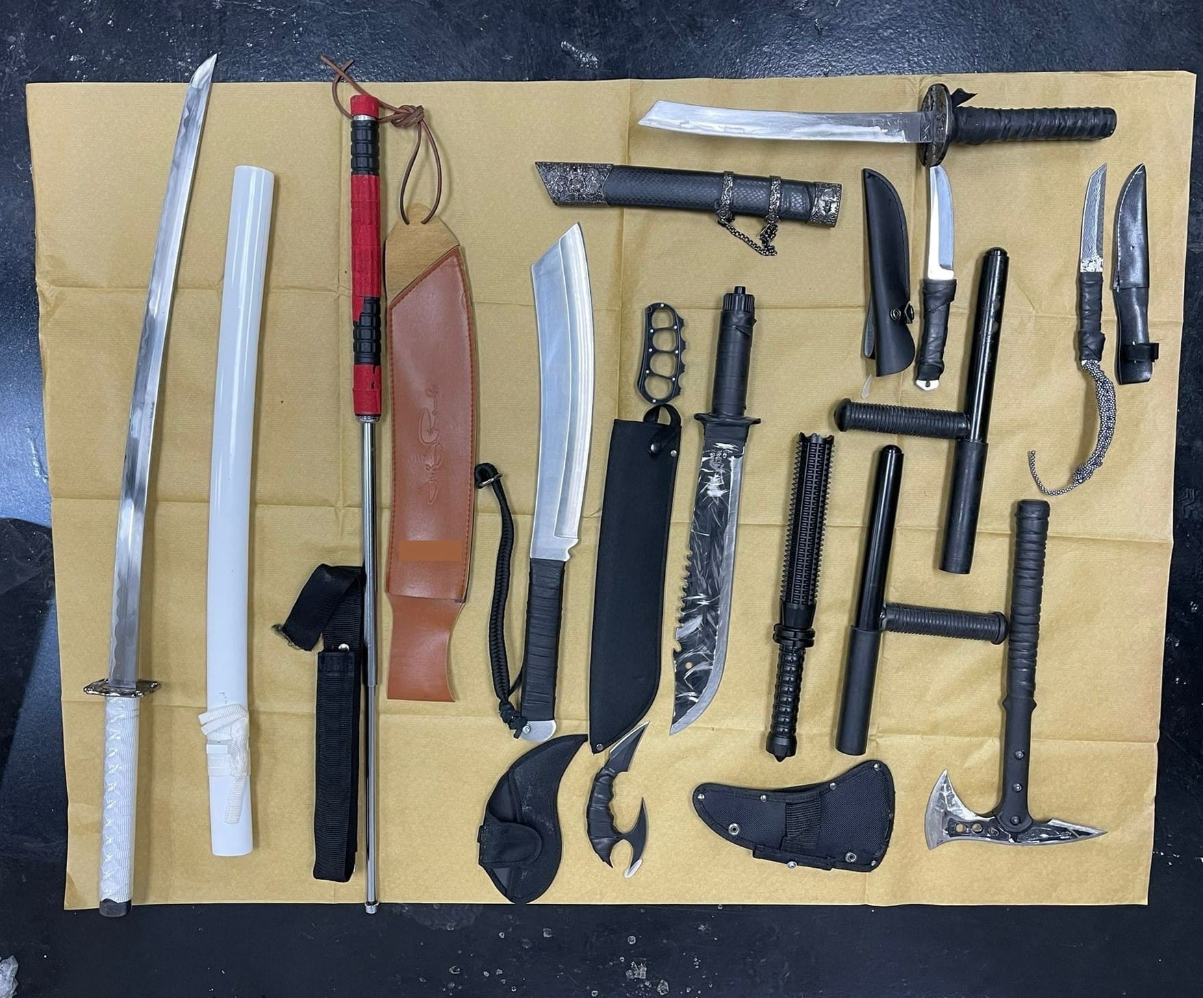 Collection of swords, knives, batons and brass knuckles laid out on a brown surface.