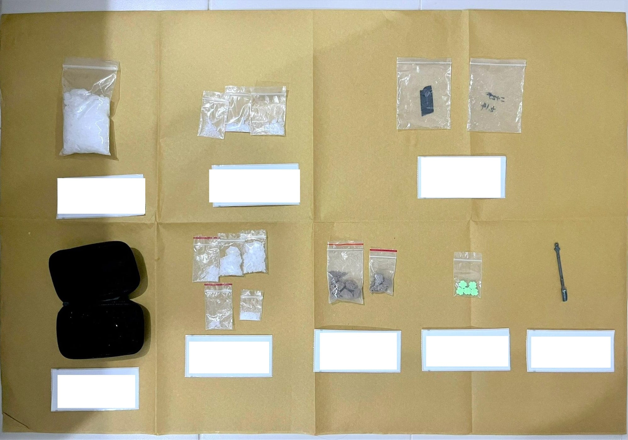 Evidence spread on brown paper: bags of white powder, green tablets, gray rocks, black case, and metal tool.