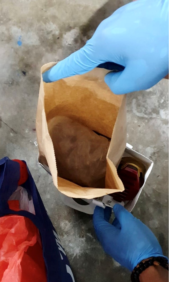 Gloved hands reach into a brown paper bag. A second white bag contains a red and gold object.