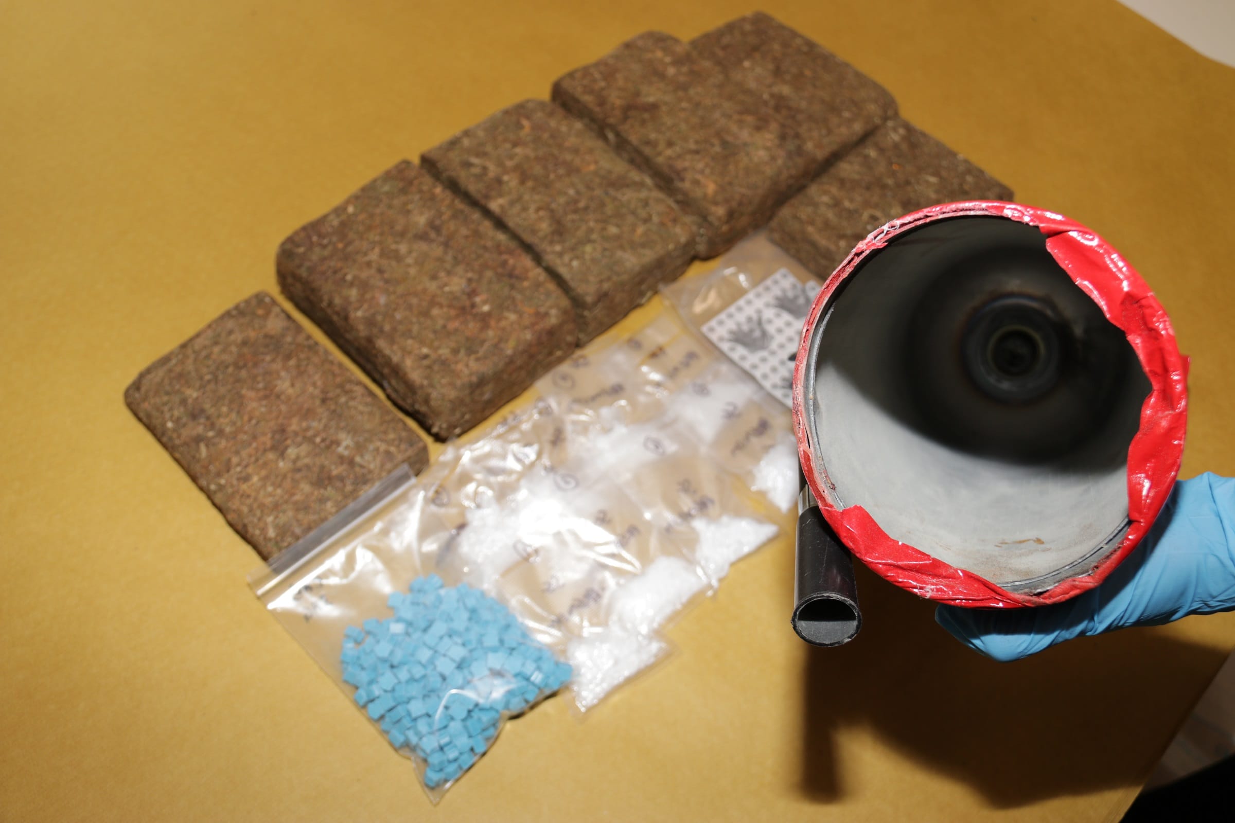 Six brick-shaped items, small bags of blue and white pills, and a gloved hand holding a red-taped metal tube.