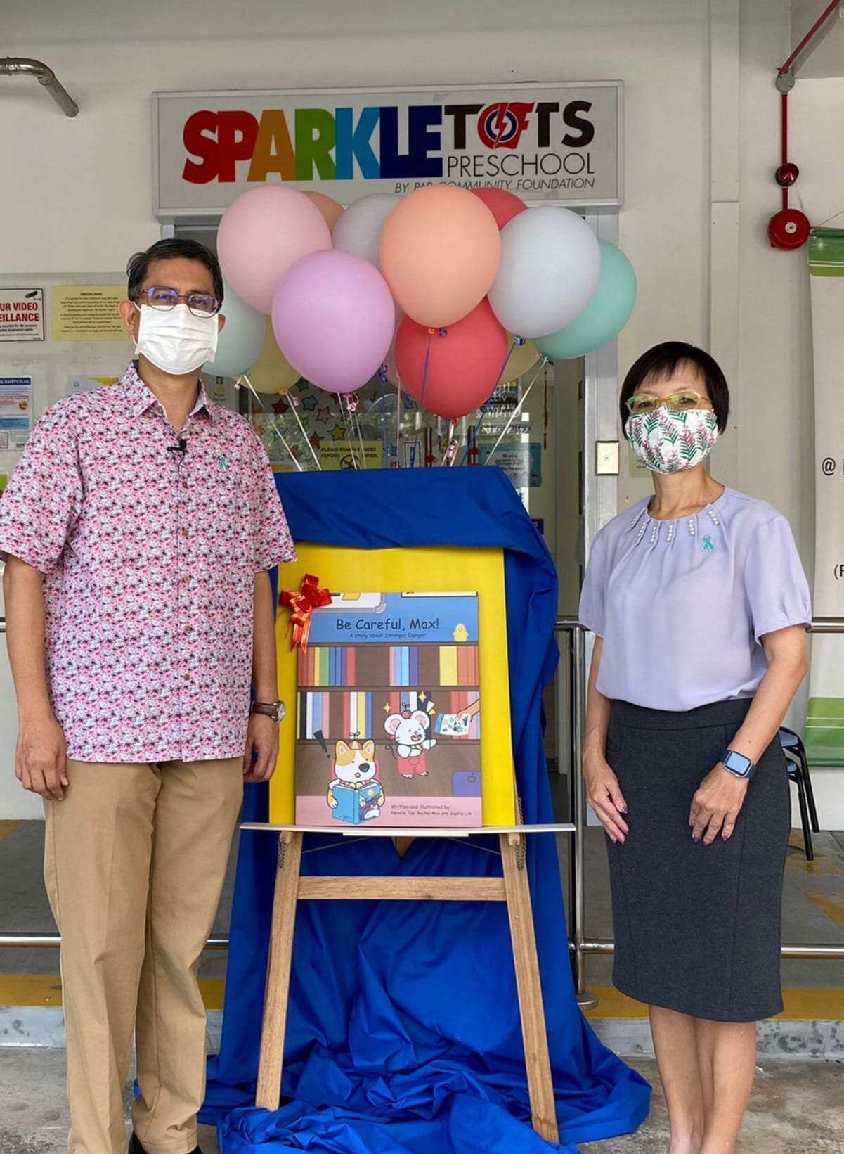 Two masked adults stand next to the book "Be Careful, Max!" at Sparkletots Preschool, with balloons behind.