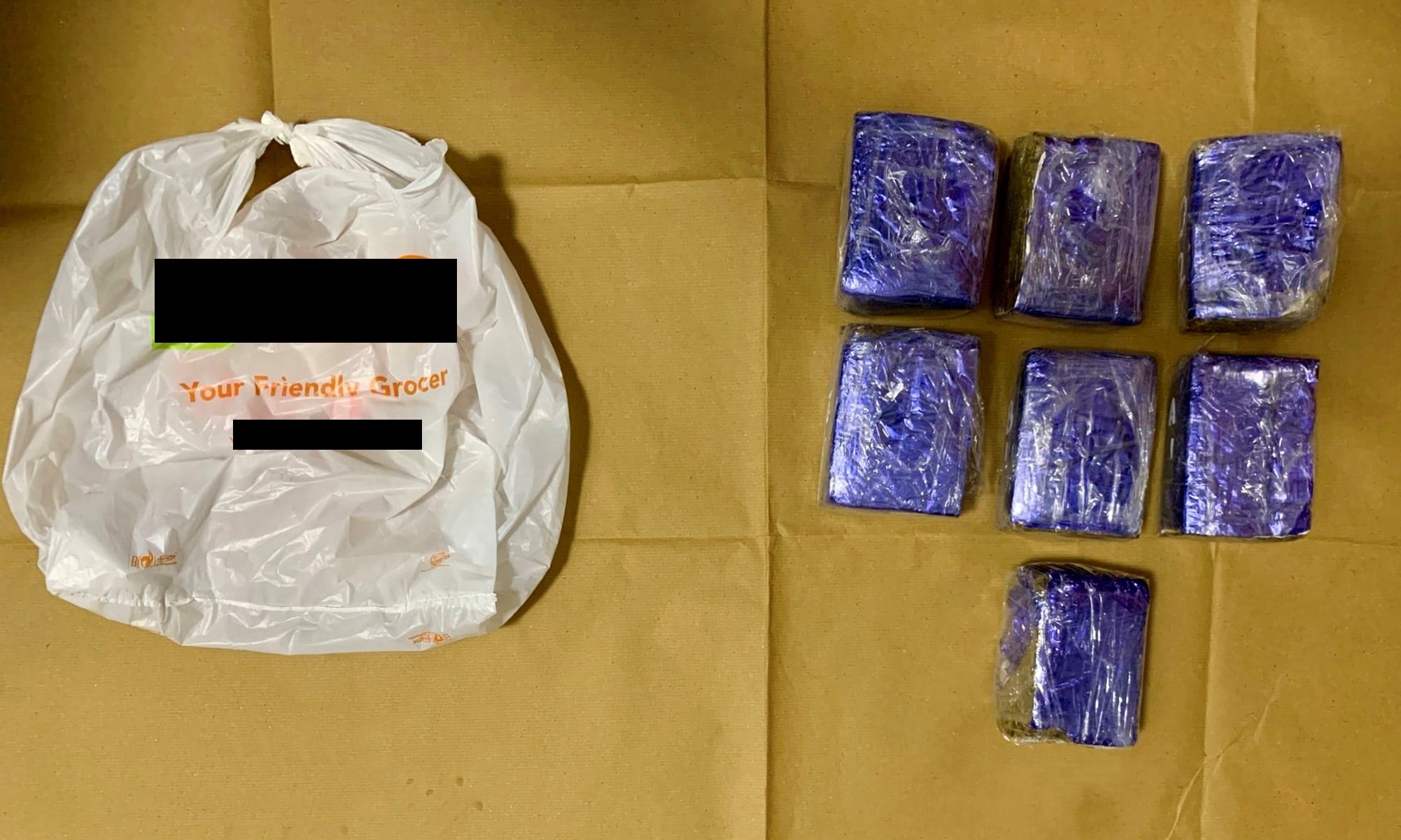 White plastic grocery bag with "Your Friendly Grocer" text, and seven blue wrapped brick-shaped packages on brown surface.