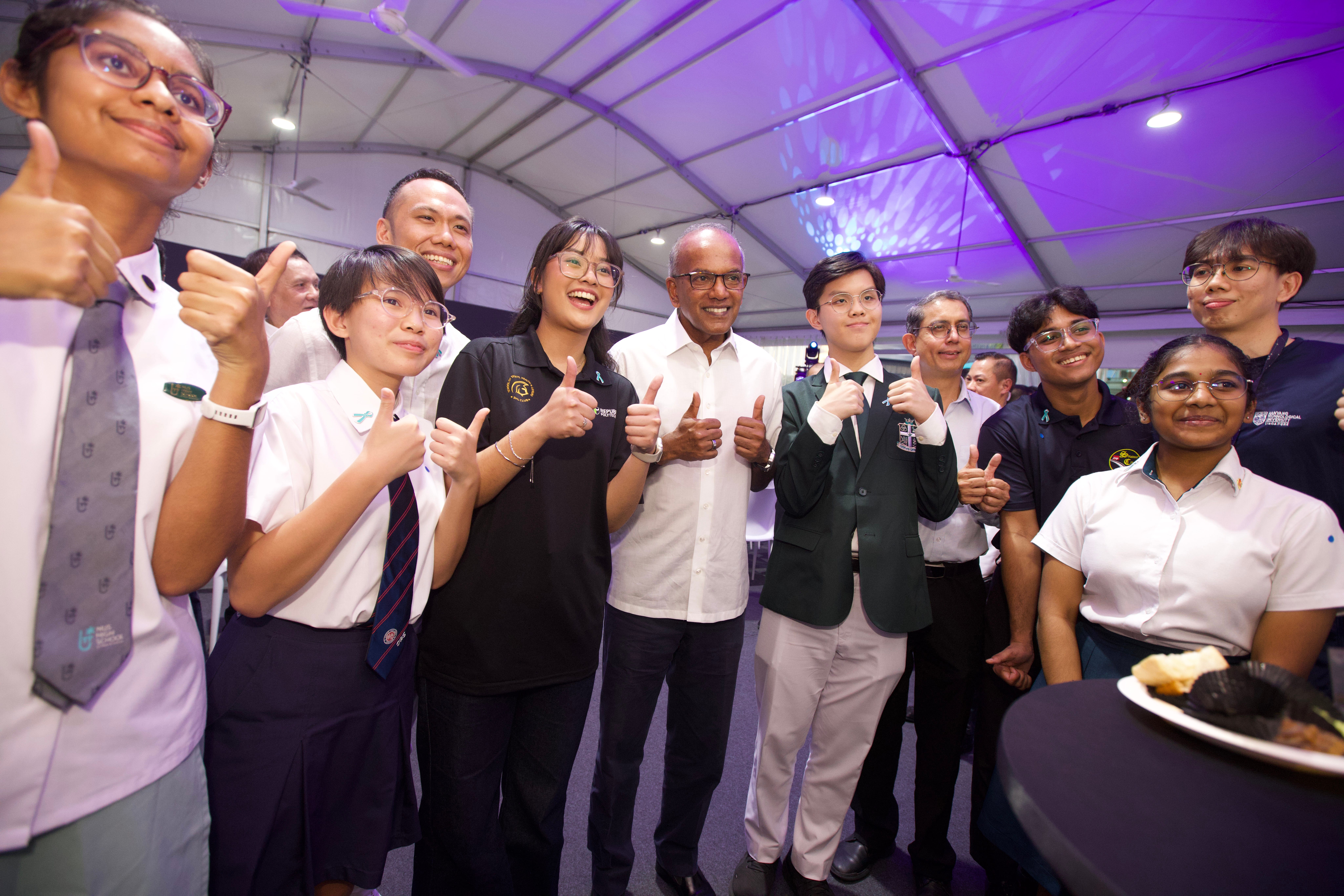 Group of students and adults giving thumbs up under a tent, wearing uniforms and business attire.