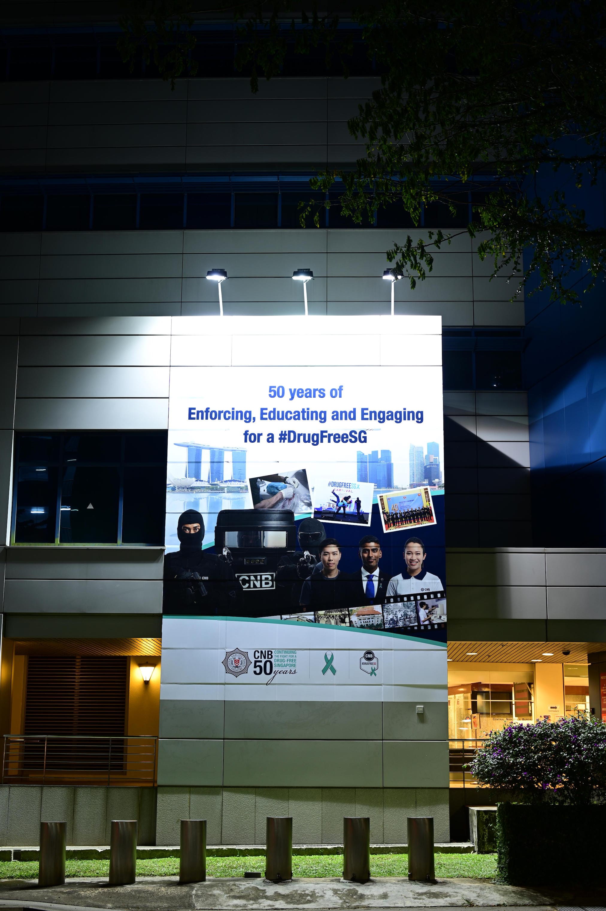 CNB anniversary banner on building: "#DrugFreeSG" with agents and Singapore landmarks.