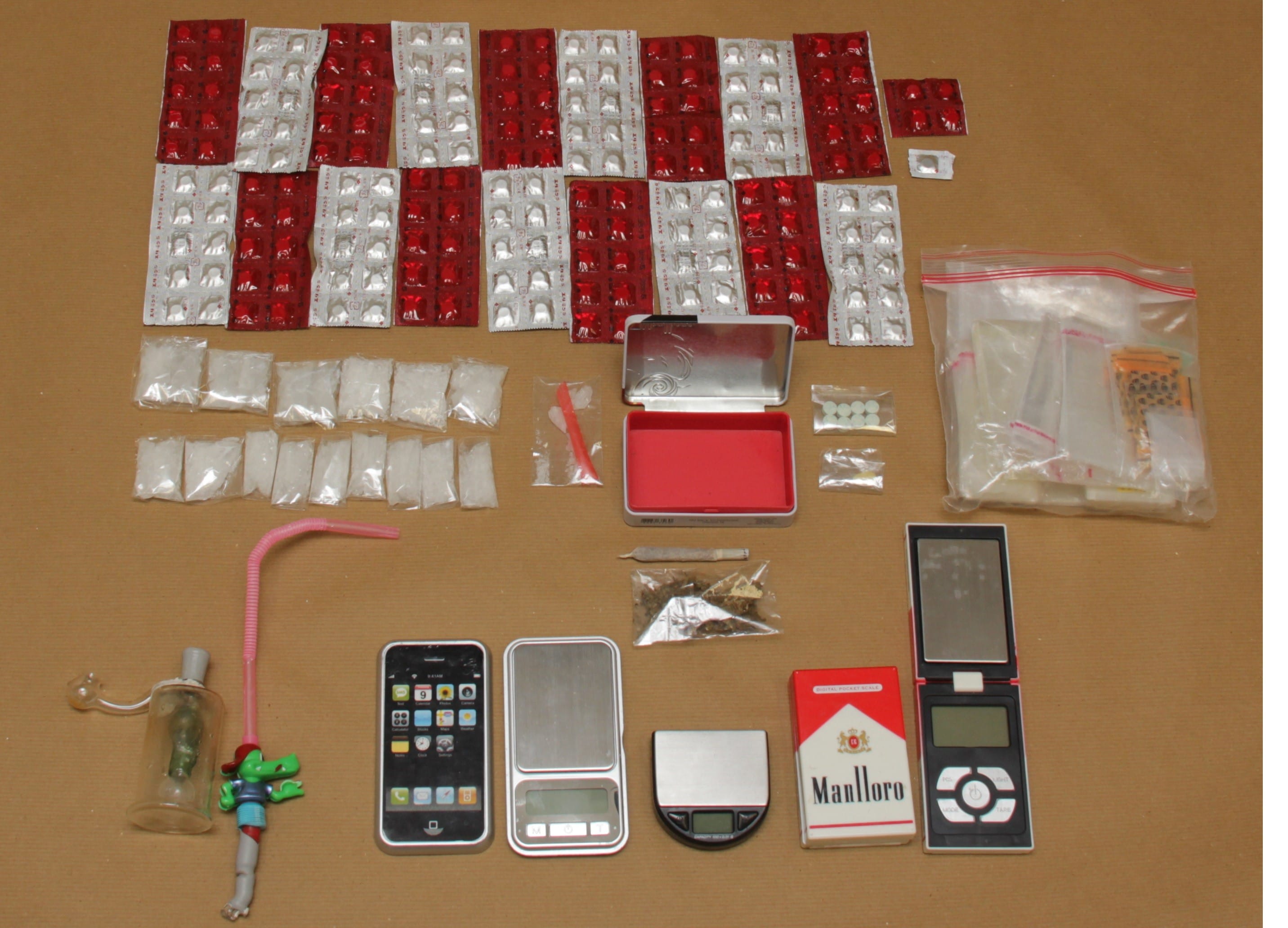 Pills in blister packs, plastic bags with white substance, phone, digital scales, "Manlloro" cigarette pack, and accessories.