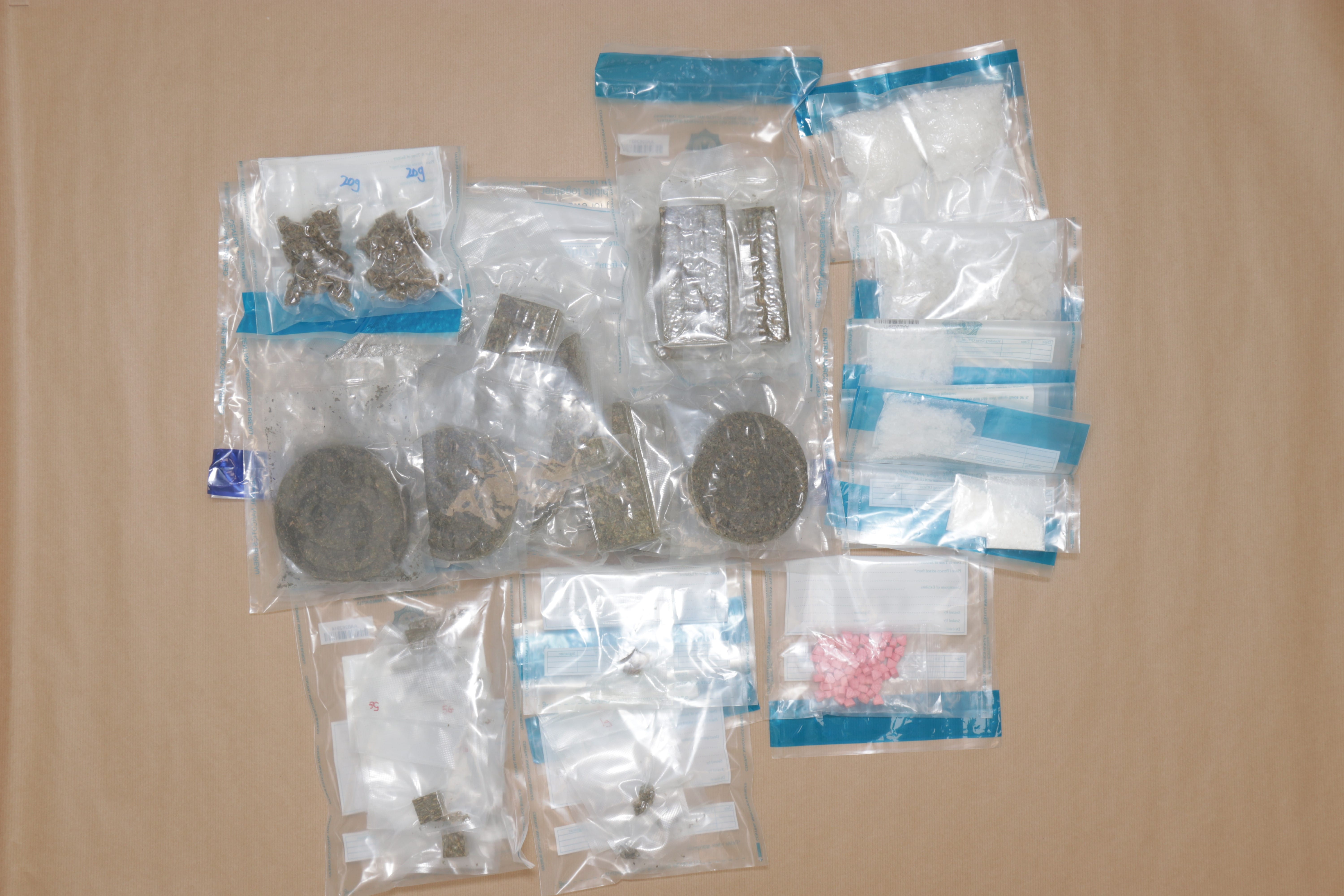 Sealed plastic bags with various powders, herbs, and pink pills arranged on a light brown surface.