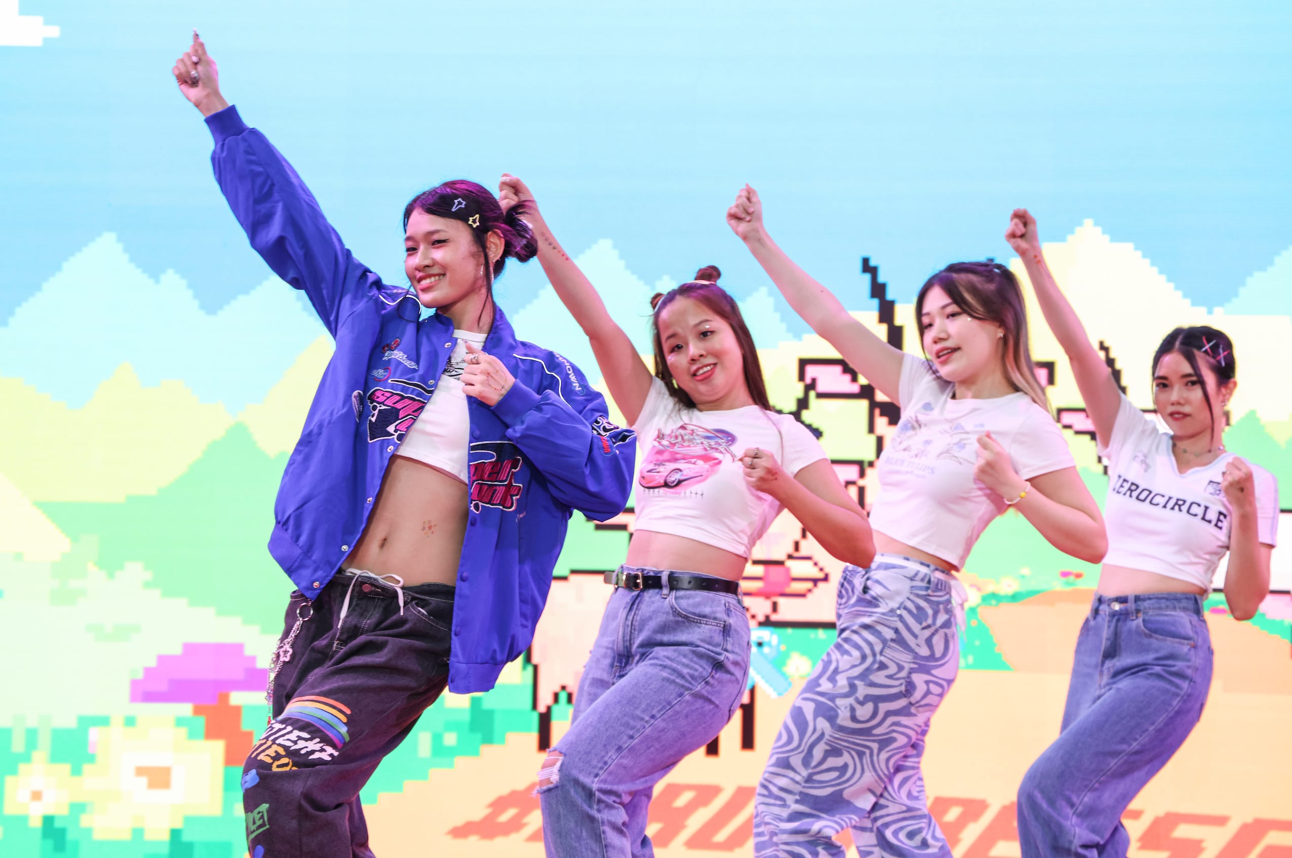 Five people in crop tops and jeans dancing in front of a pixelated backdrop.