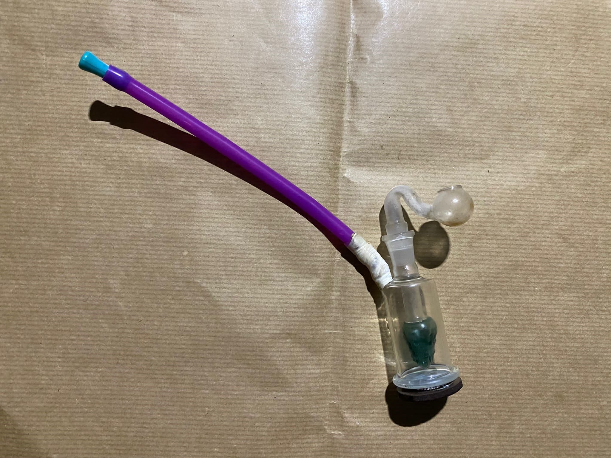 Glass pipe with green substance and purple stem, lying on brown paper.