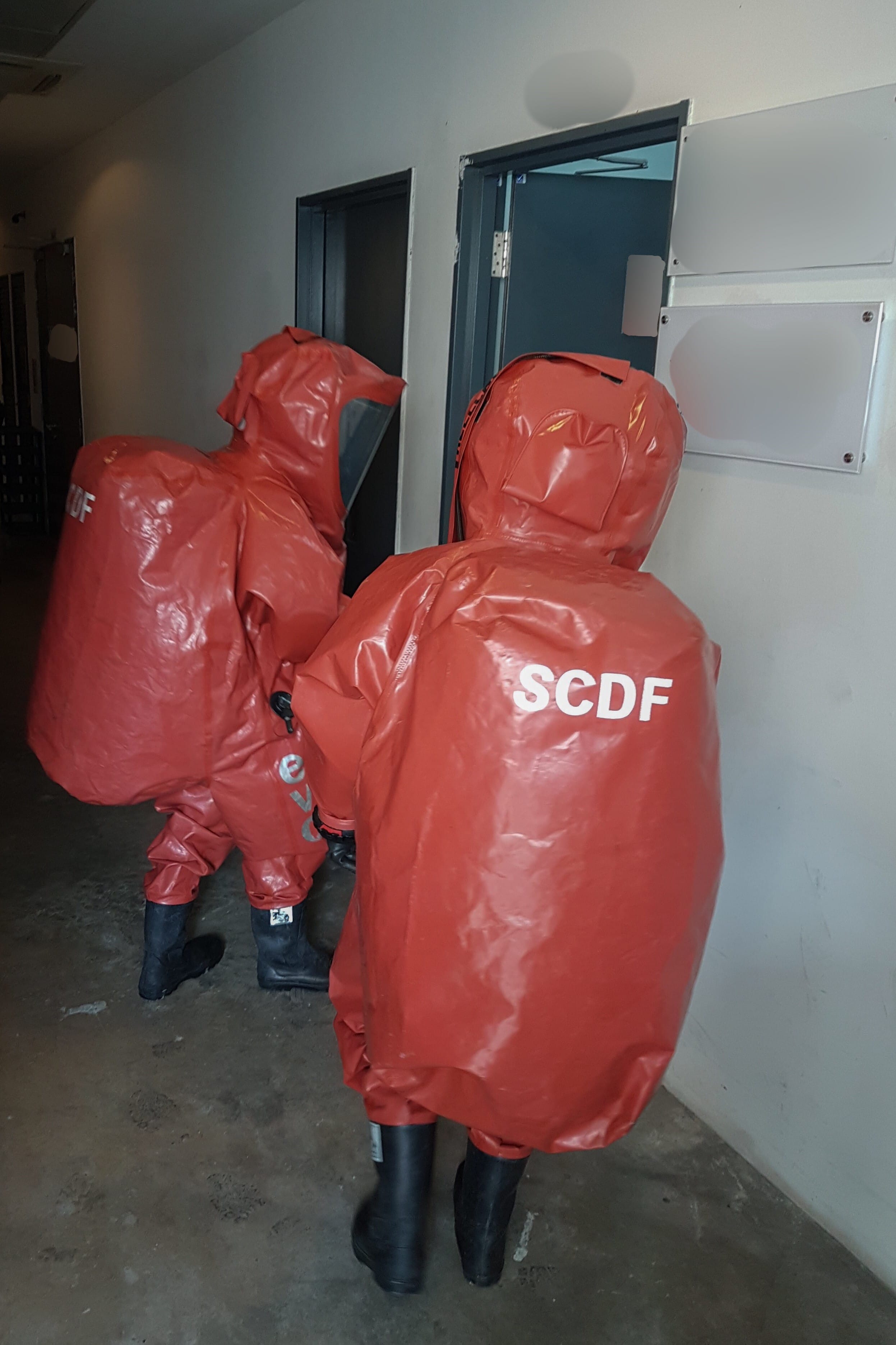 Two SCDF personnel in red hazmat suits and black boots walk in a hallway.
