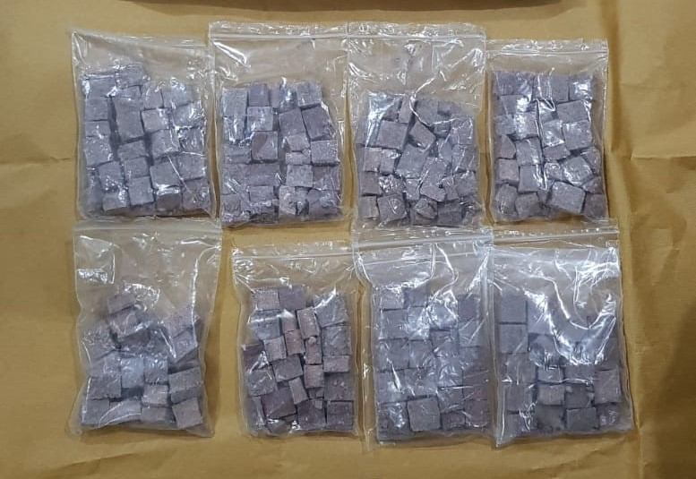 Eight clear plastic bags filled with small, purple cube-shaped items on a tan surface.