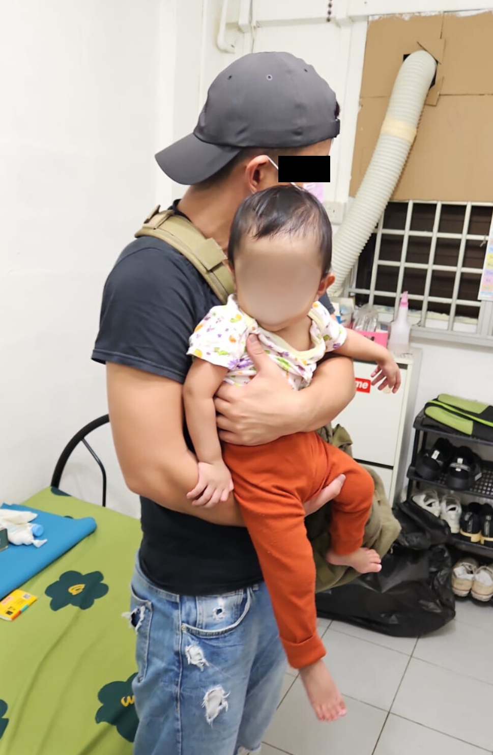 Person in hat holds baby in patterned shirt & orange pants; bed, AC setup, shoe rack visible.