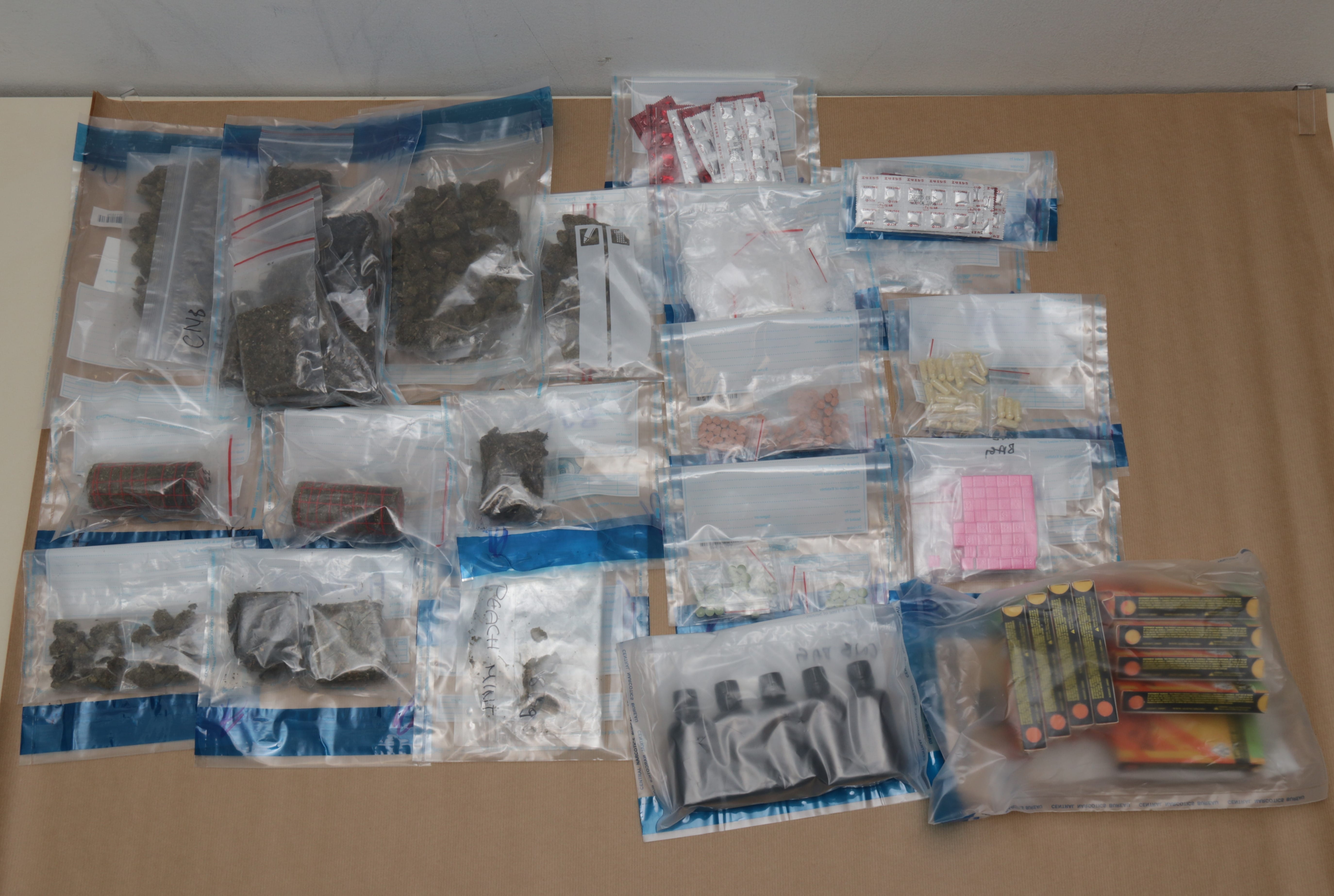 Multiple clear bags containing substances: green buds, pills, capsules, and packaged materials.