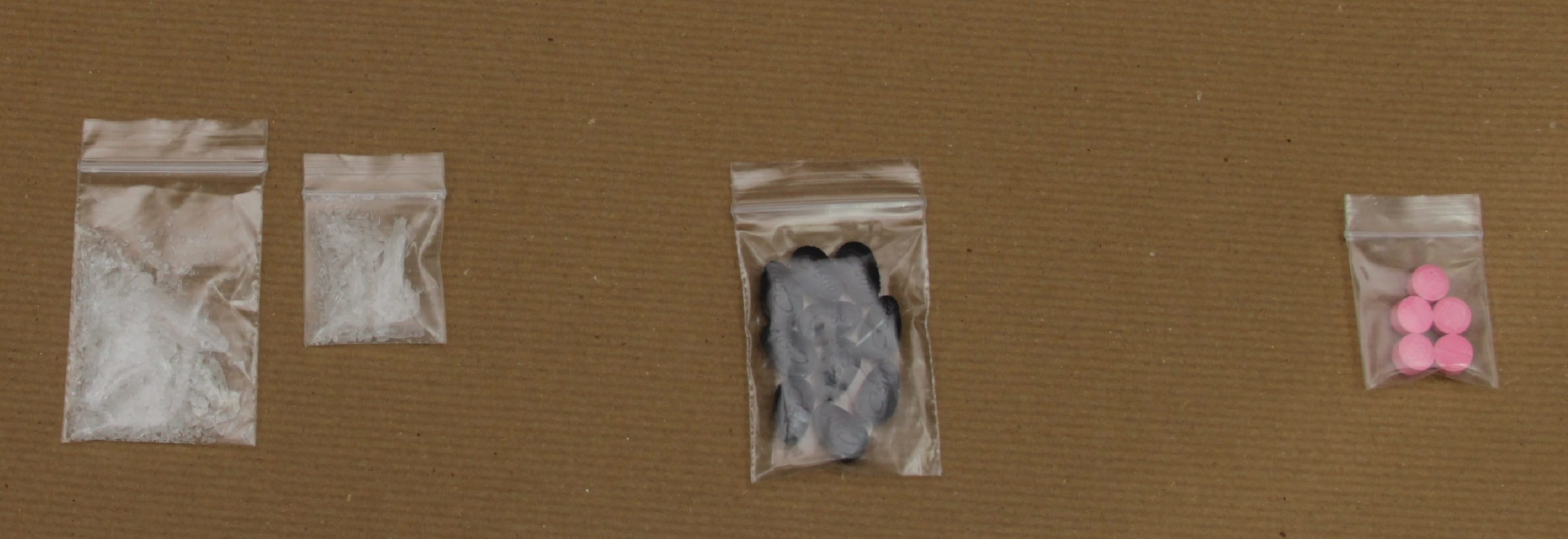 Four clear plastic bags on a brown surface contain white crystals, gray pills, and pink pills.