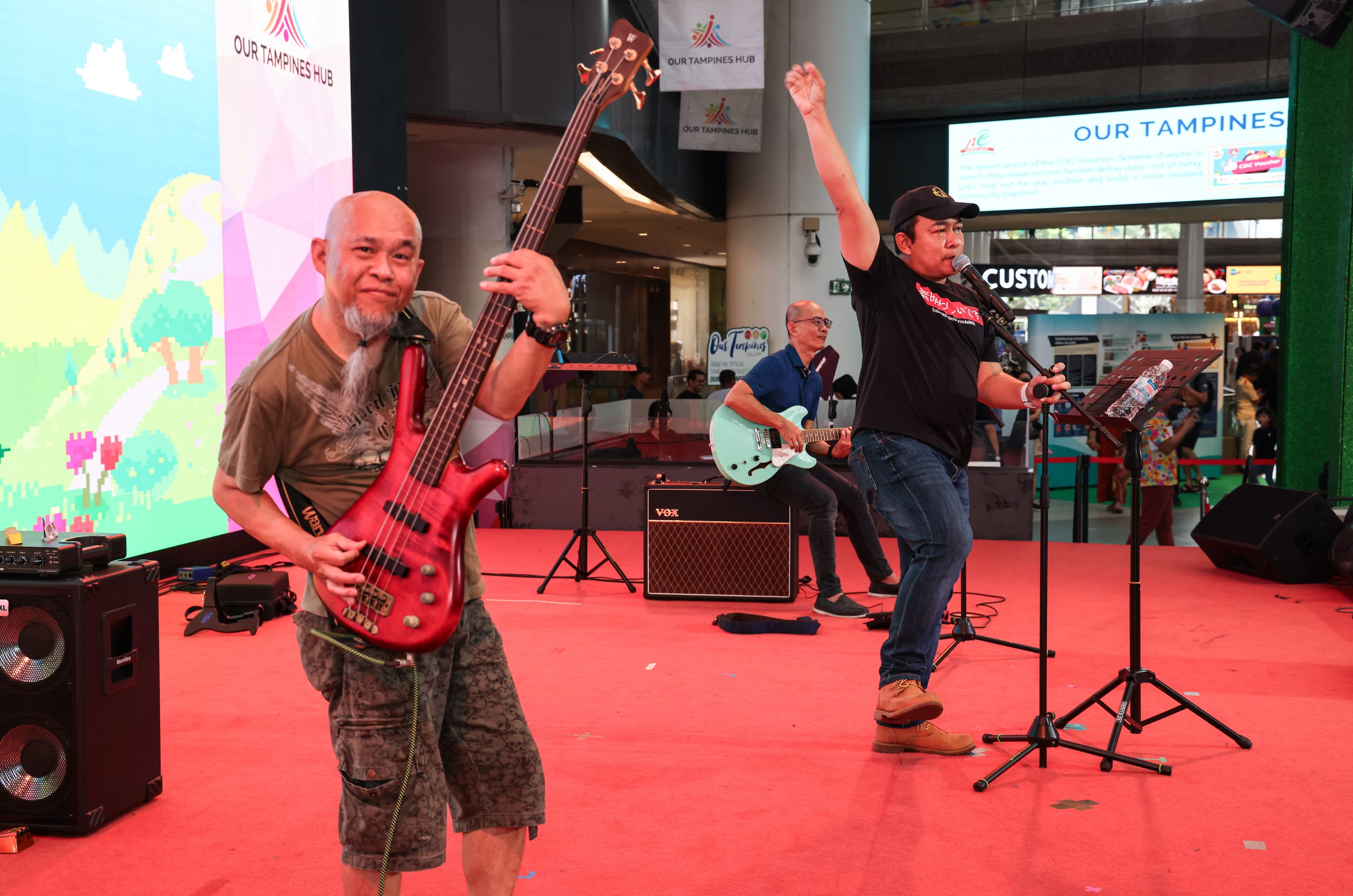 Band performing live on stage. A singer and two guitarists, with "Our Tampines Hub" banners and signs.