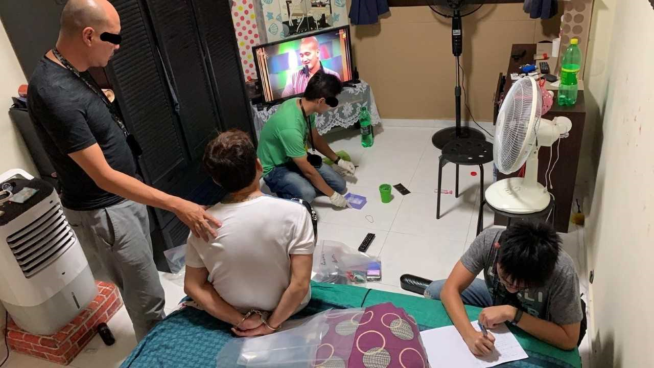 Interior with three figures; one in handcuffs on a bed, two figures searching and documenting. TV visible in background.