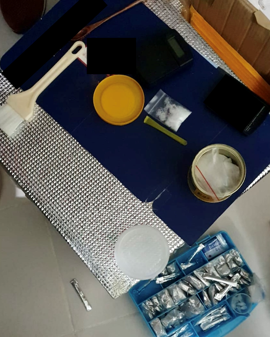 Overhead shot of materials: containers, powders, scale, and a brush on metallic/blue surfaces.