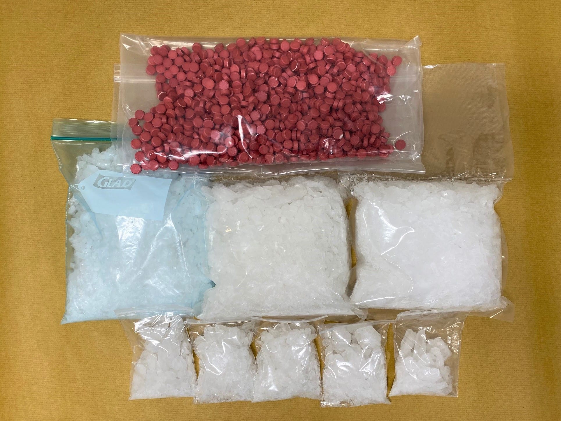 Nine sealed bags, one with red pills, the others with white crystals, on a tan surface. One bag labeled "Glad".