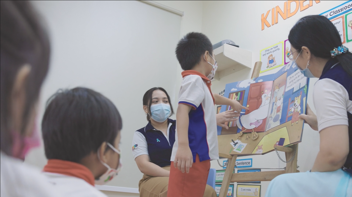 Boy pointing at book on easel in classroom; teacher looks on. Everyone wears masks.