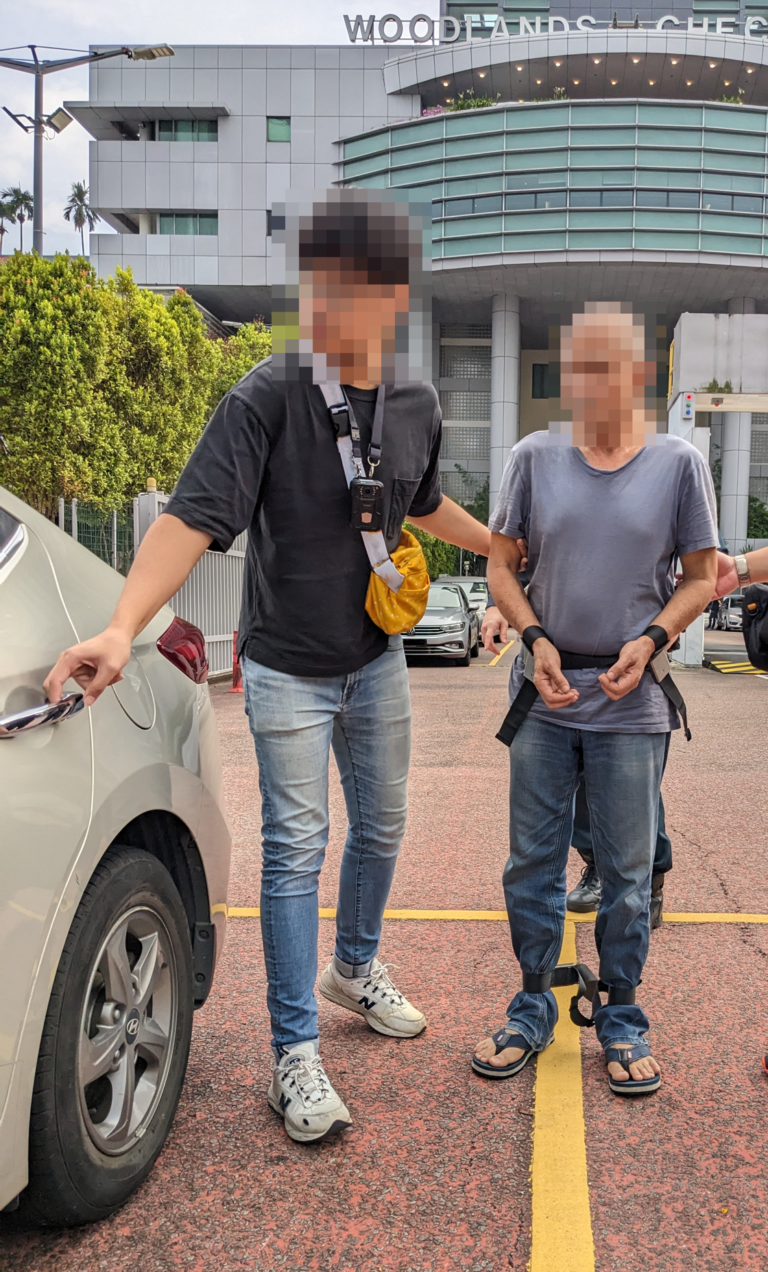 Man with restraints escorted to car by another, Woodlands Checkpoint building in background.