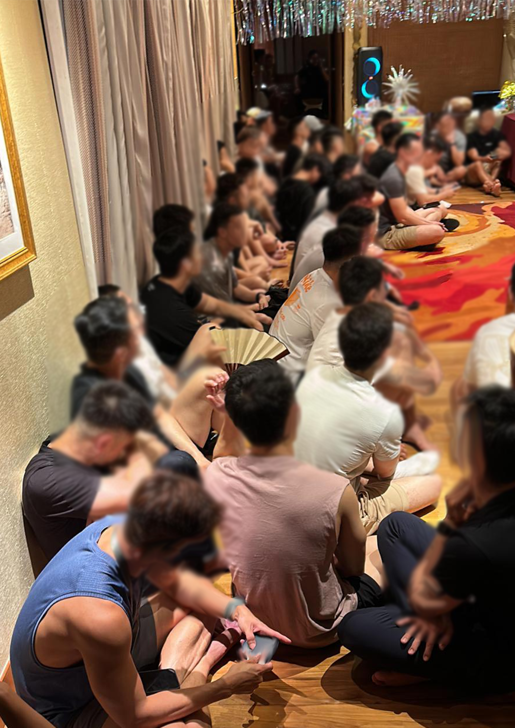 Many blurred figures sit cross-legged on a carpeted floor in a room with decorations.