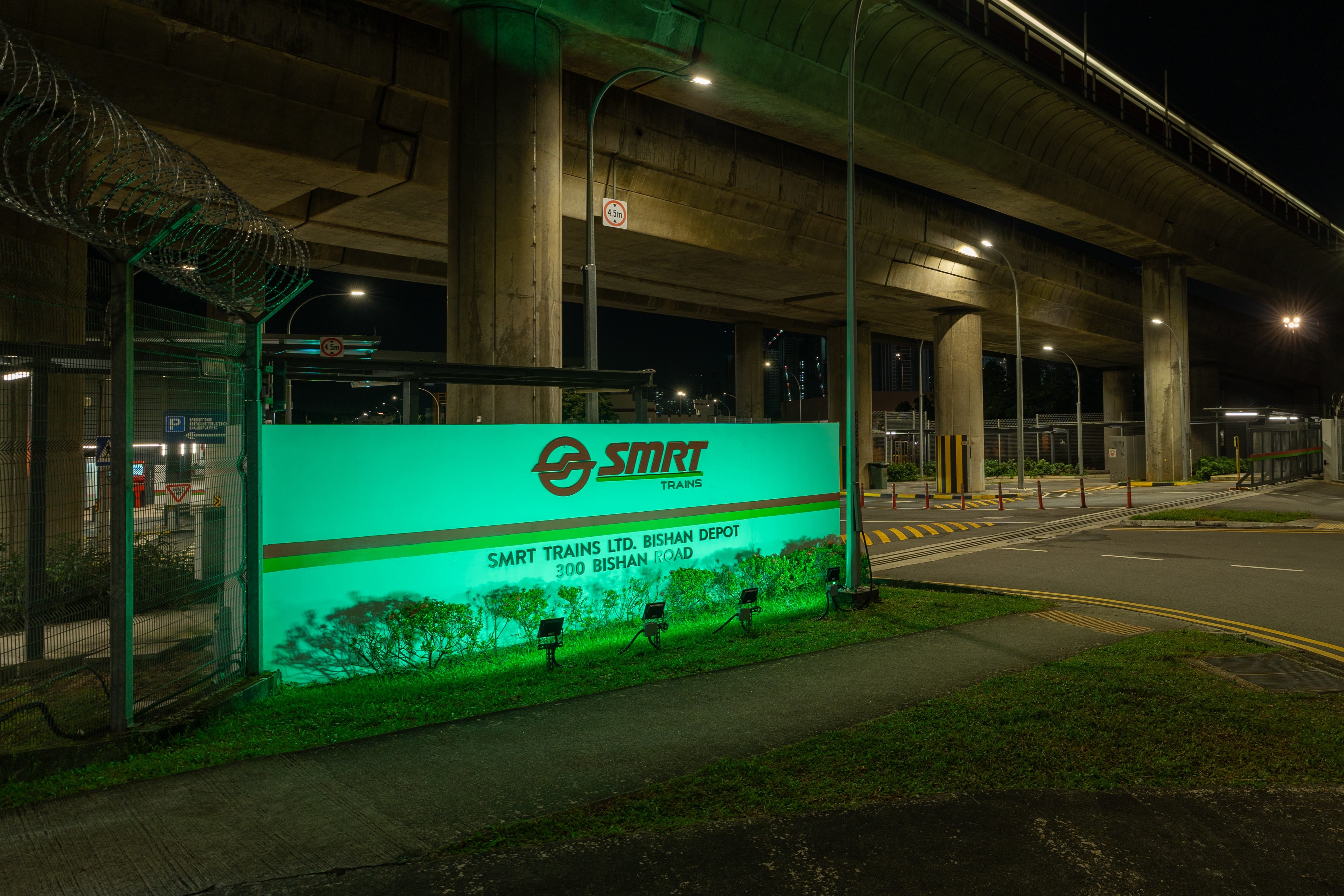 Illuminated SMRT trains sign at night, under a highway overpass, next to fenced area.