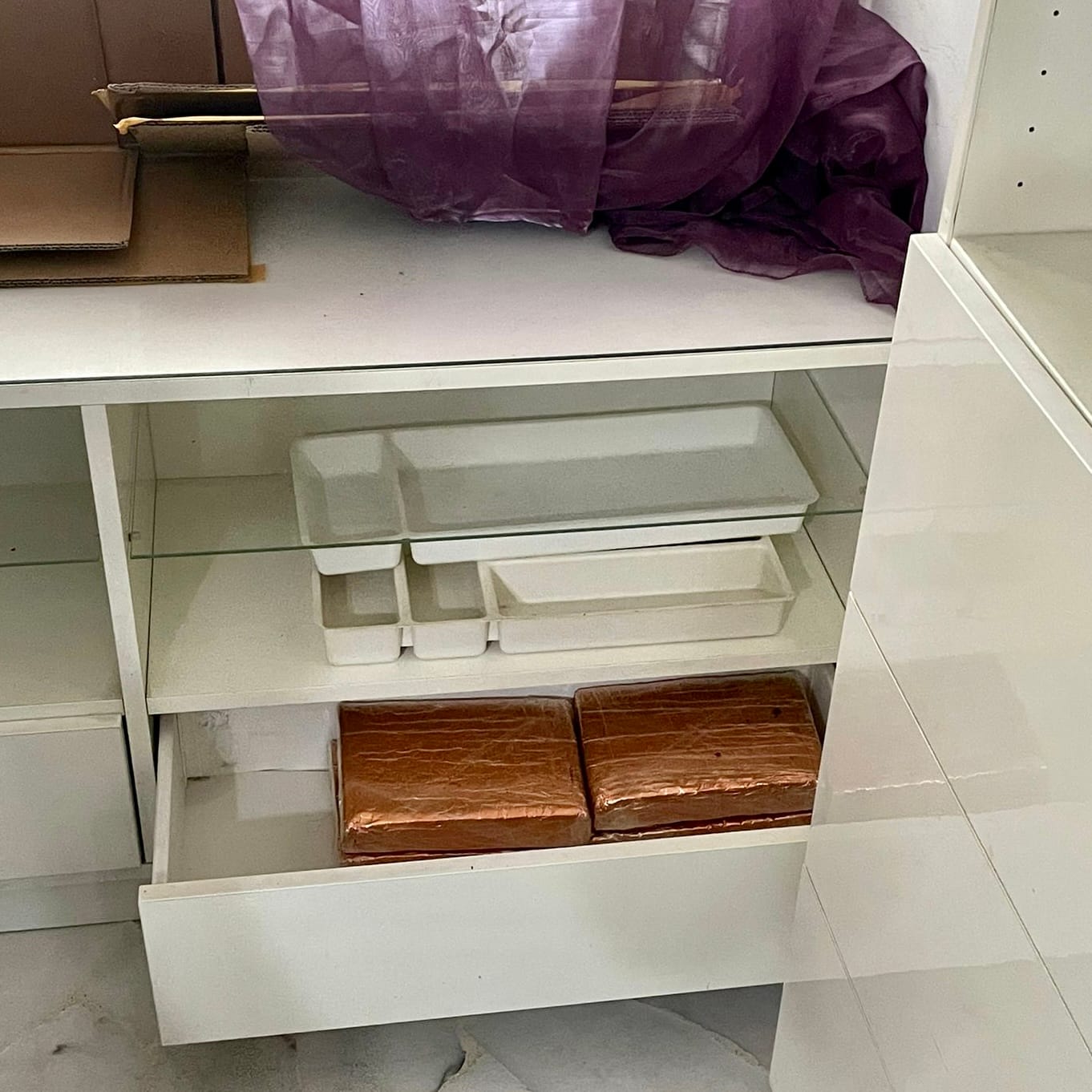 White cabinet with open drawer; two wrapped brown loaves inside. Top shelf: boxes and purple fabric.