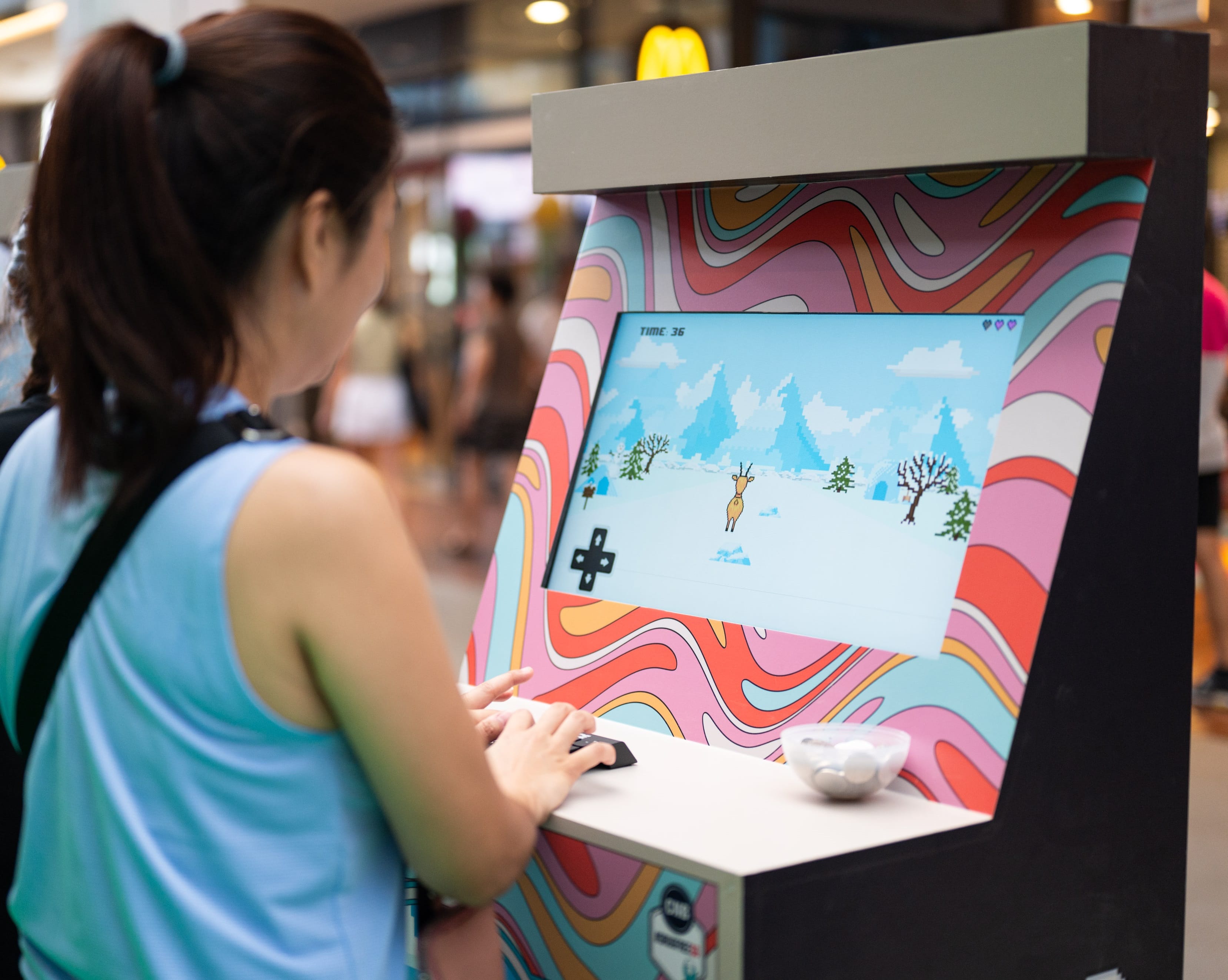 Person playing retro game on a psychedelic patterned arcade machine. Pixelated reindeer on screen.