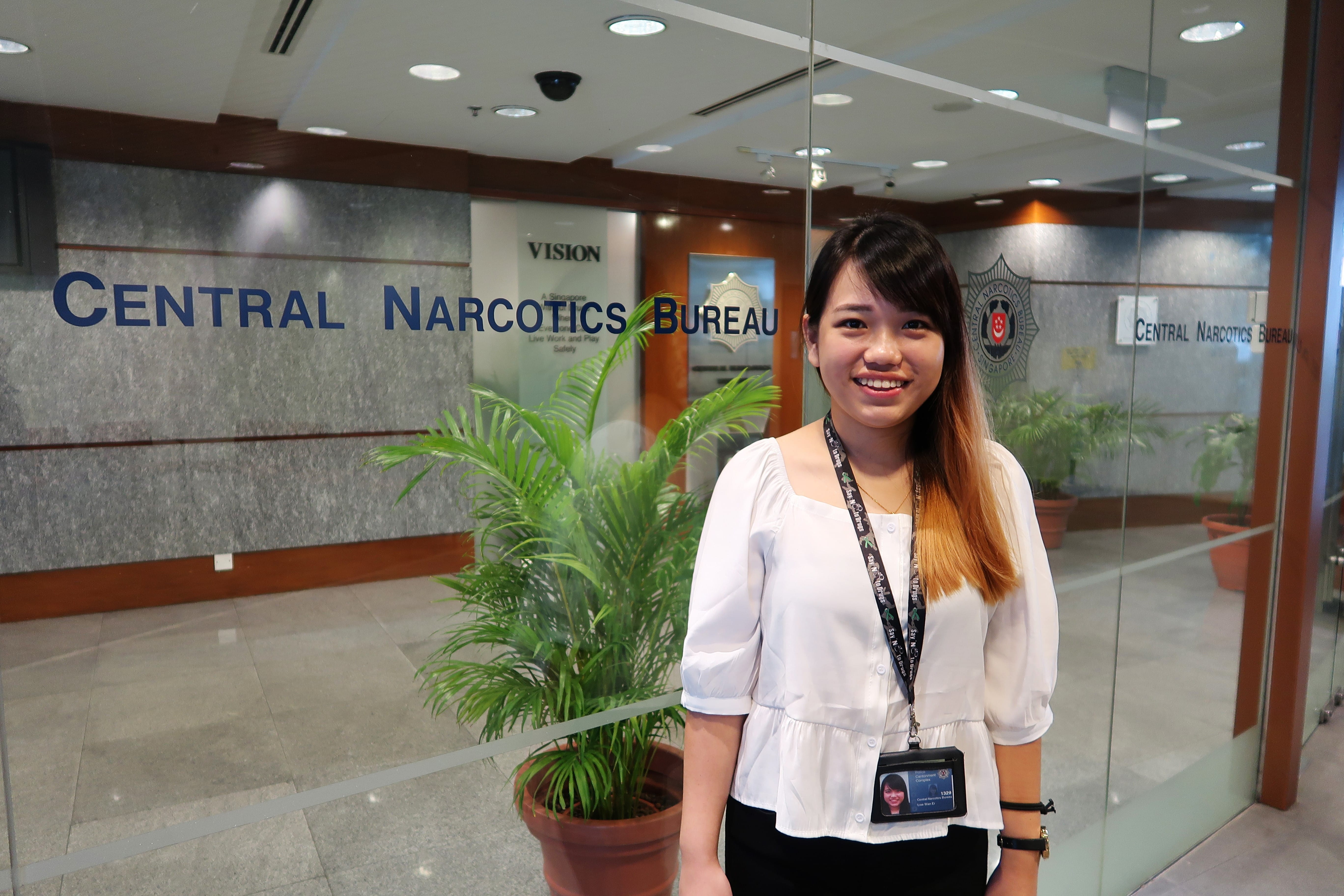 Woman in white blouse standing inside Central Narcotics Bureau office, smiling at the camera.