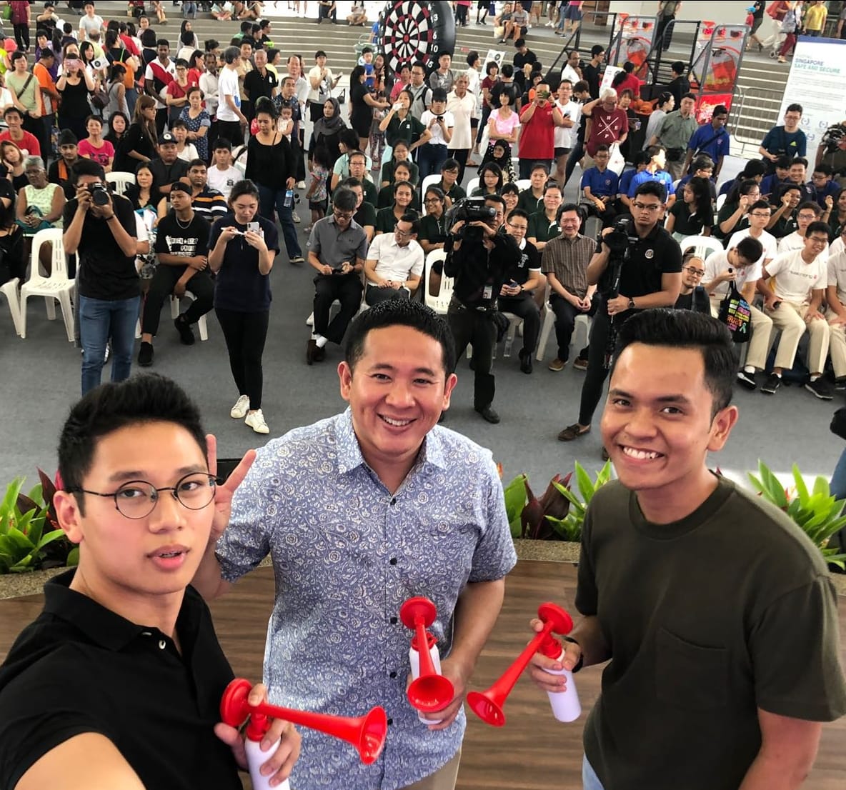 Three men hold red air horns, smiling at camera. Large crowd visible behind on steps.